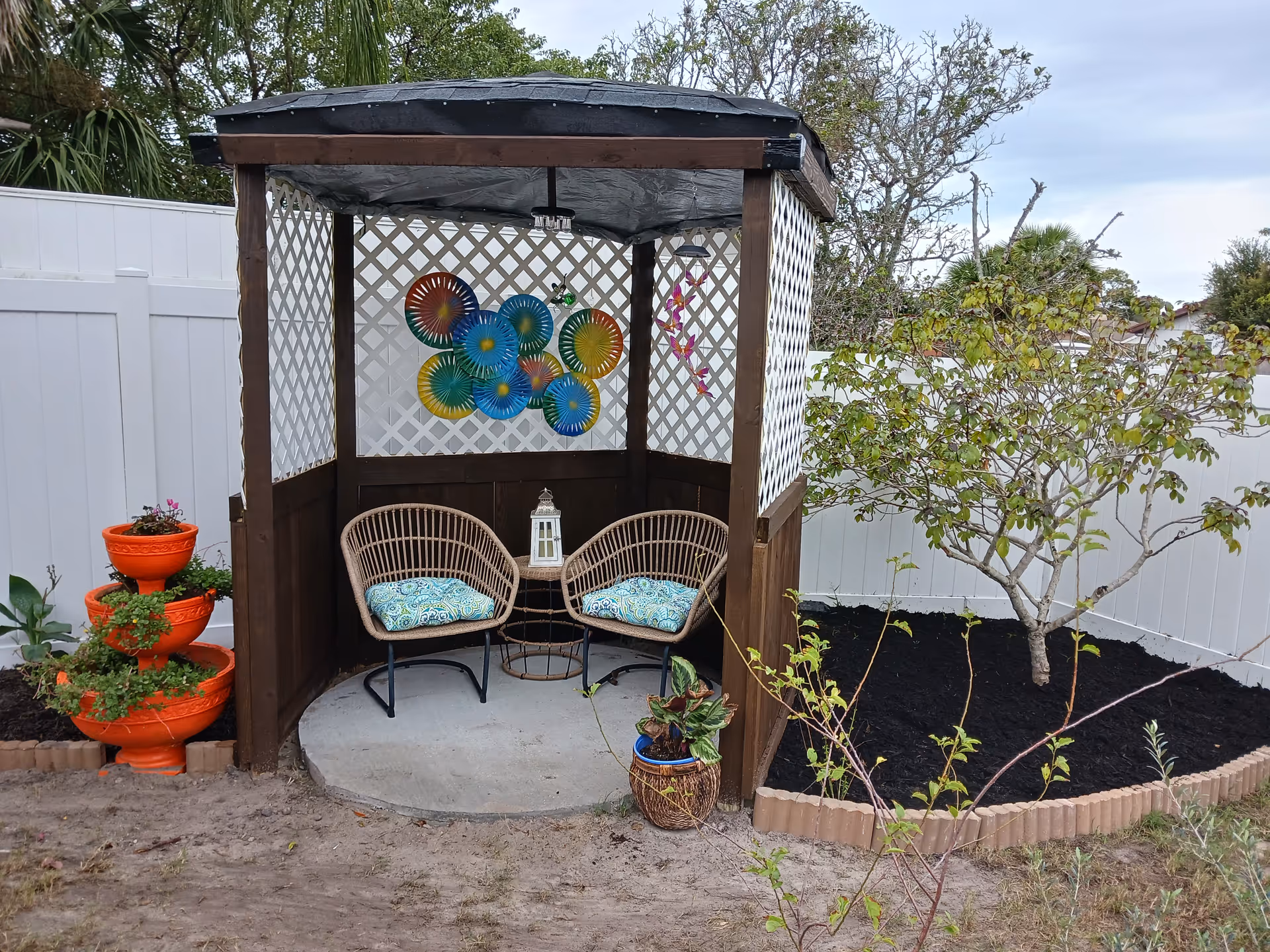 Outdoor wooden lattice gazebo with two wicker chairs, cushions, lantern, and garden plants.