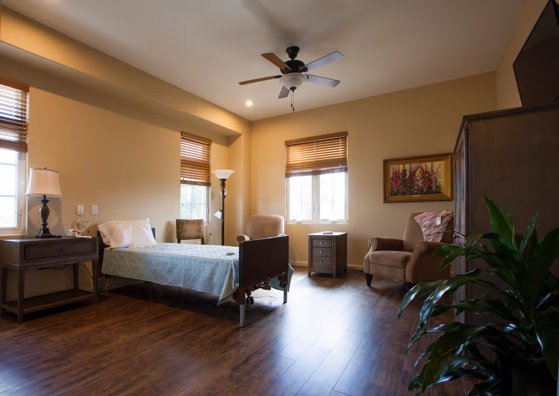 Spacious bedroom in a senior care facility with a bed, nightstand, chairs, and plants.