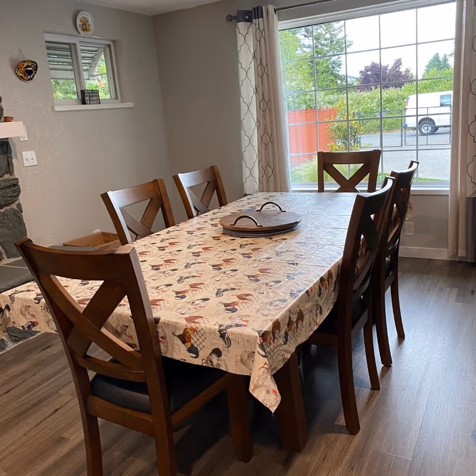 A dining room with a long table, chairs, patterned tablecloth, and large windows.