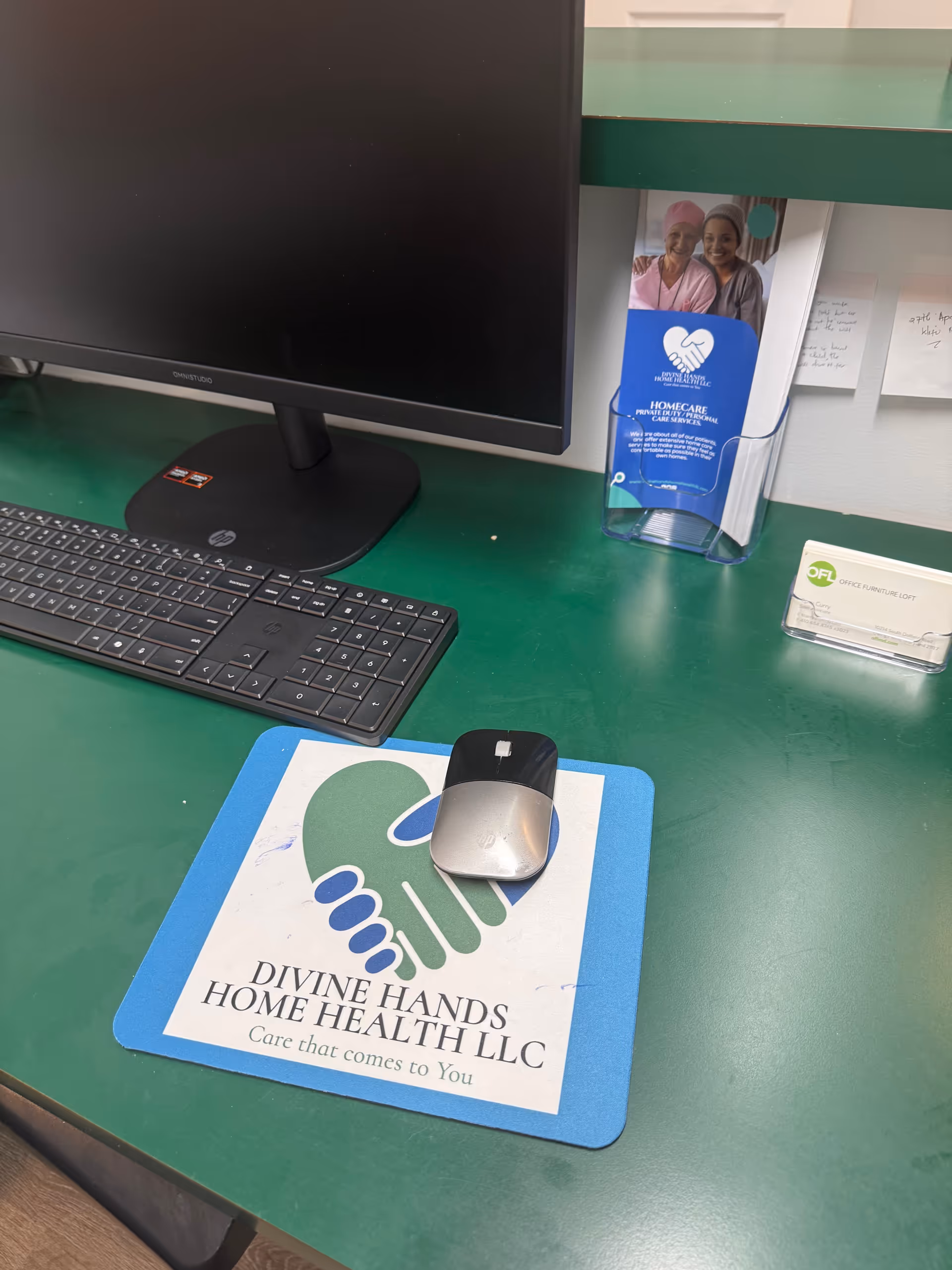 Desk with computer setup and a Divine Hands Home Health LLC logo mat.