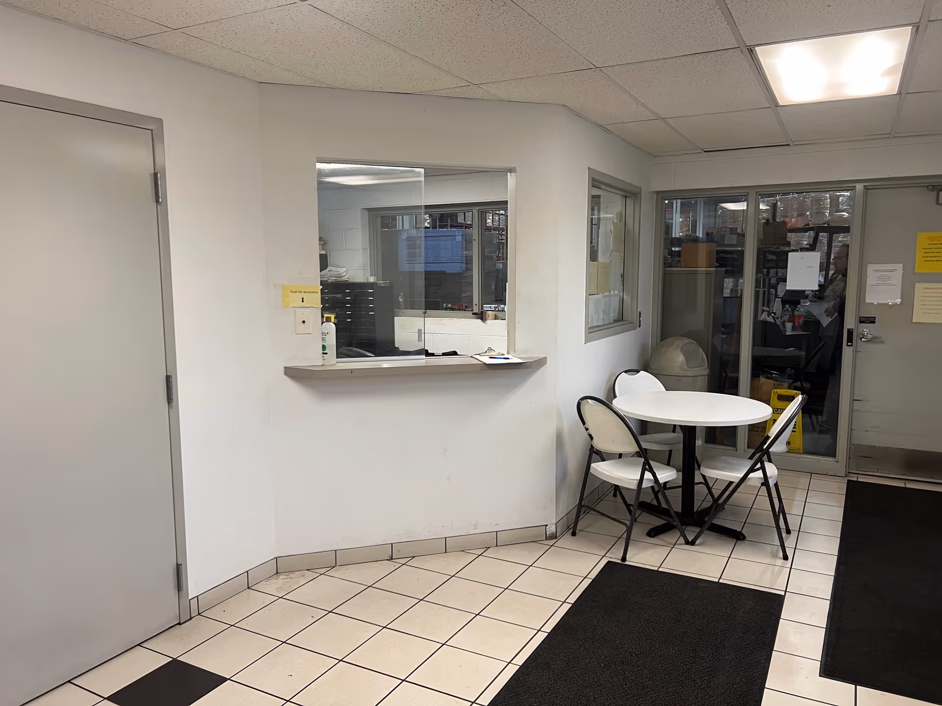 Reception window with counter and small round table with white chairs in a tiled interior hallway.