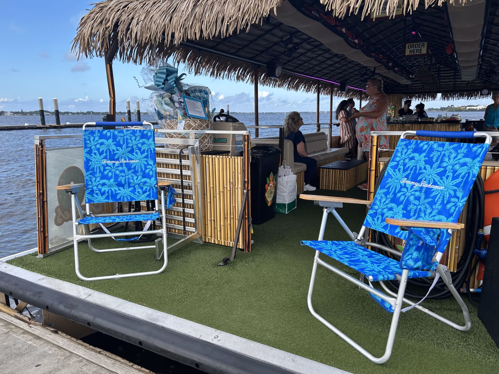 Open-air bar boat with blue palm-pattern chairs, thatched roof, and patrons inside.