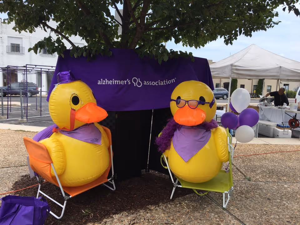 Outdoors, two yellow inflatable ducks sit beside a purple Alzheimer's Association banner.