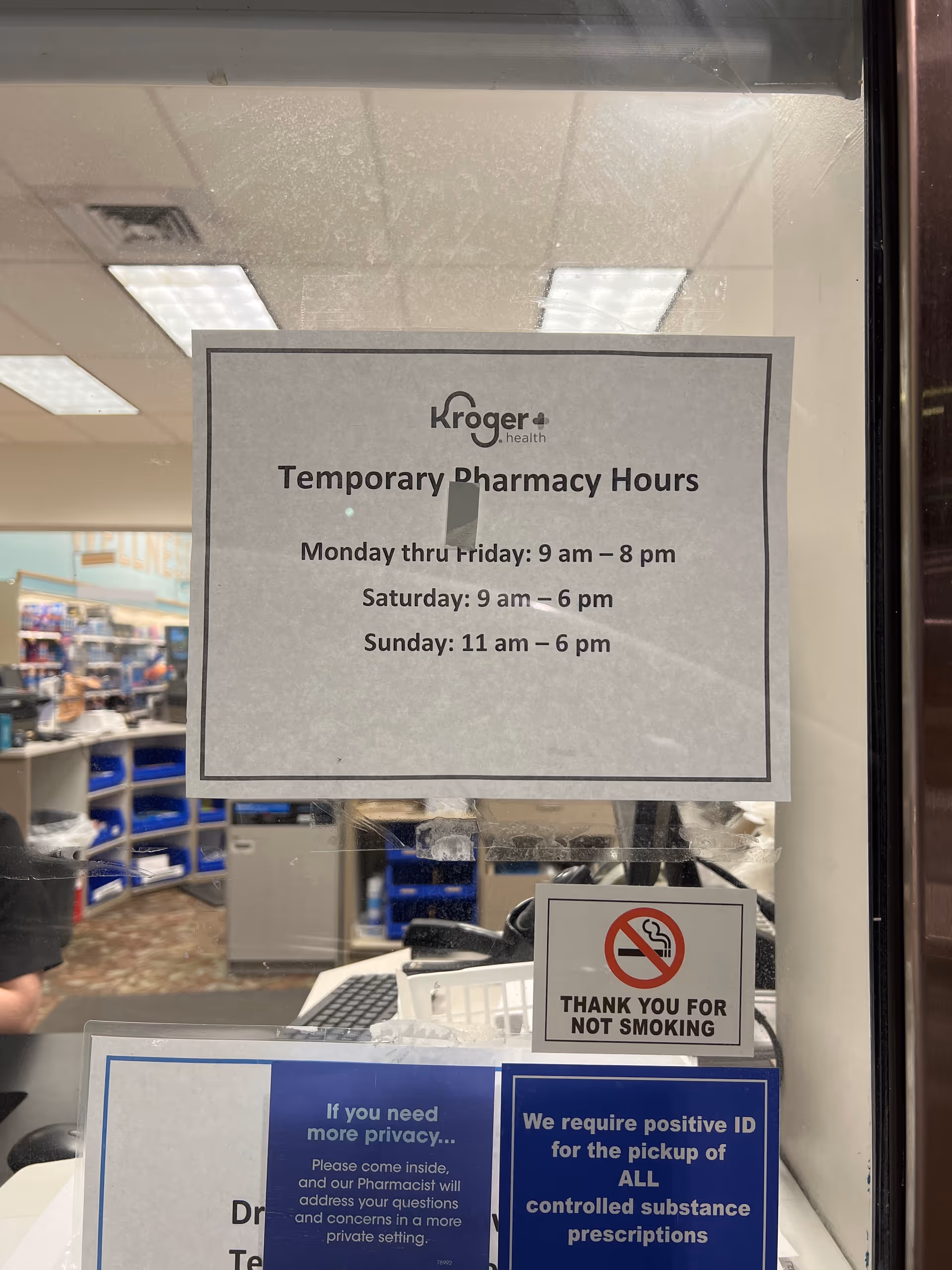 Pharmacy counter inside a facility with a sign listing temporary Kroger Pharmacy hours and shelves.