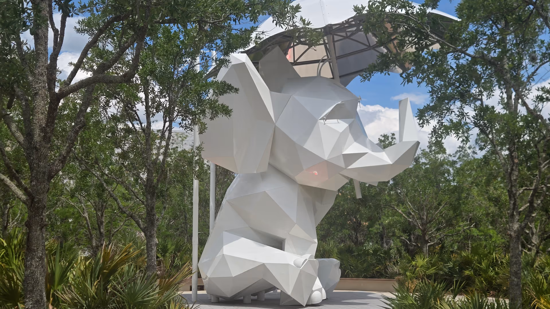 White geometric rhinoceros sculpture outdoors among trees and shrubs.