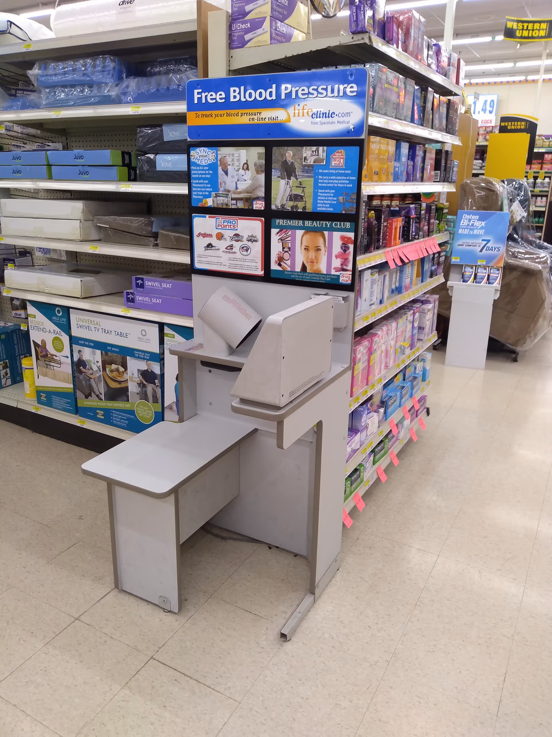 Inside a Discount Drug Mart store, a blood pressure kiosk sits beside shelves.