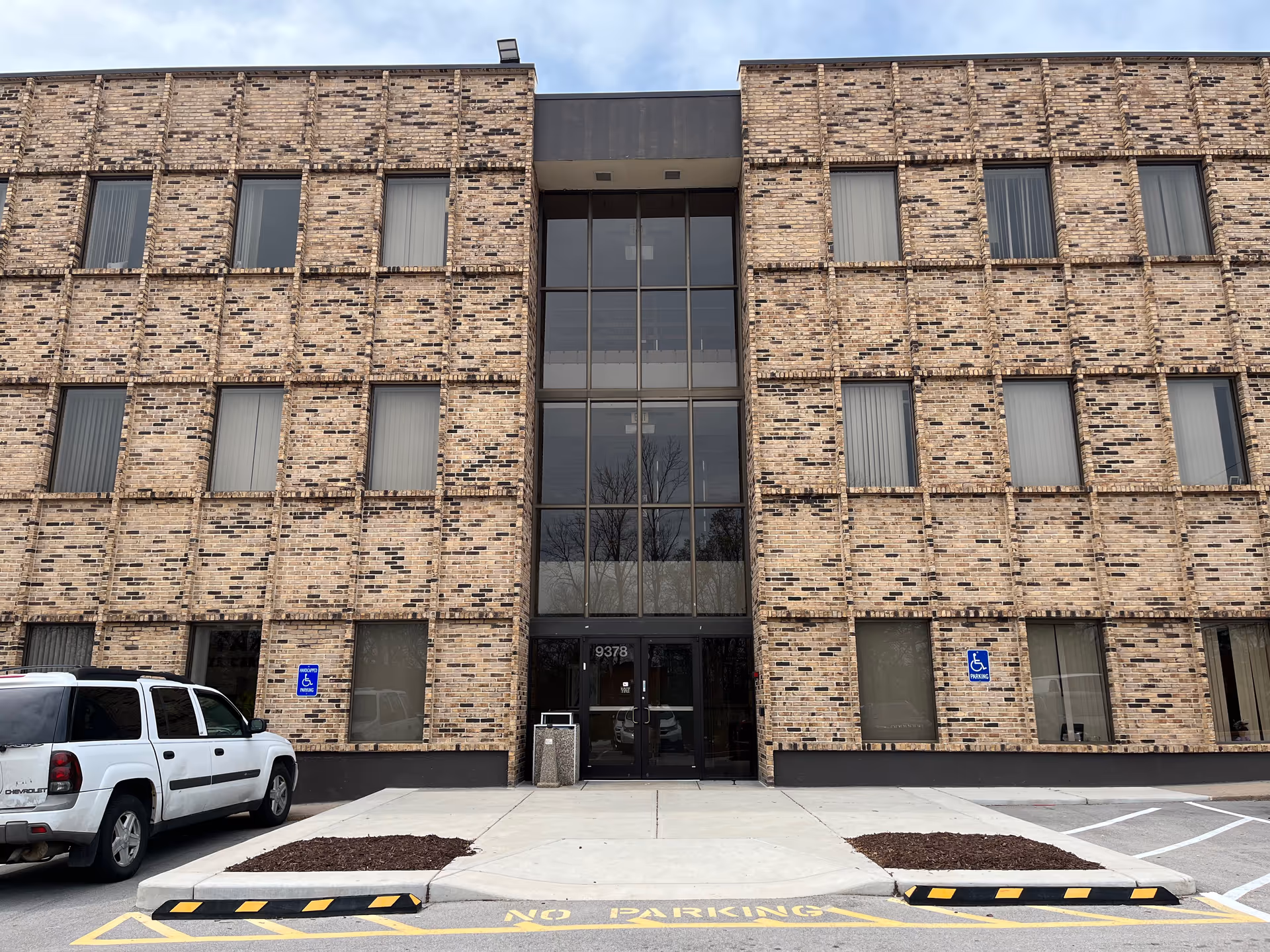 Front view of a brick multi-story facility with central glass entrance and parking.