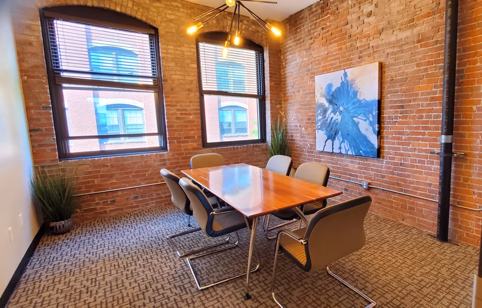Brick-walled interior conference room with a wooden table, six chairs, plants, and wall art.