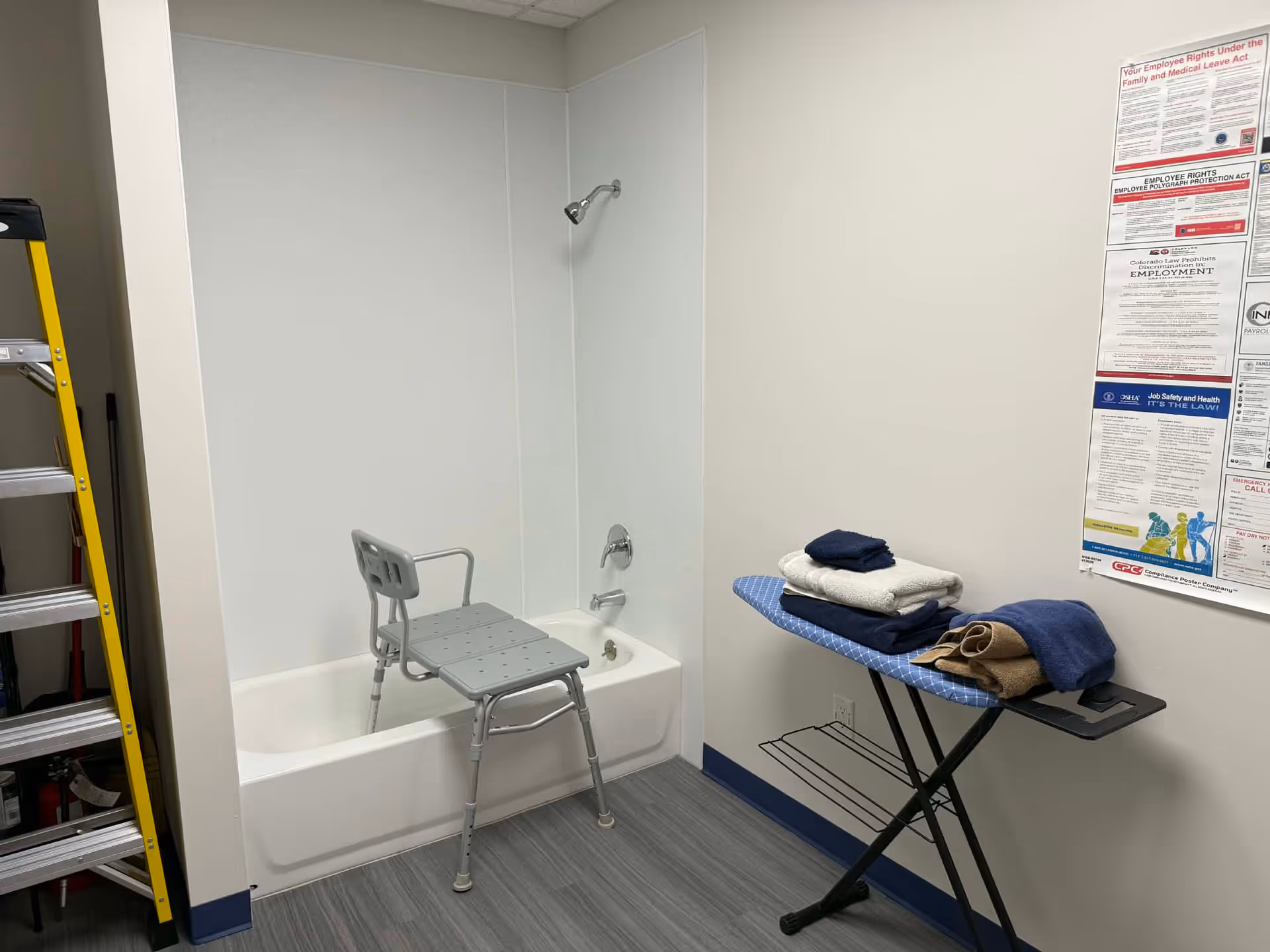 Small accessible bathroom with tub, shower chair, ironing board, and stacked towels.