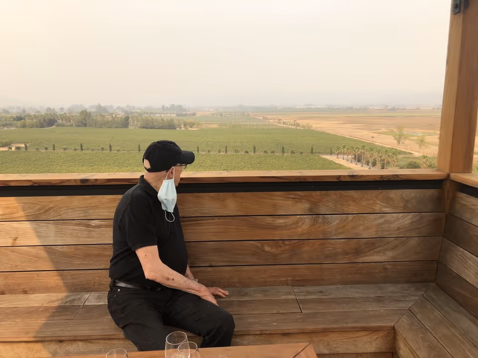 Older man sits on a wooden outdoor deck, wearing a mask, overlooking farmland.