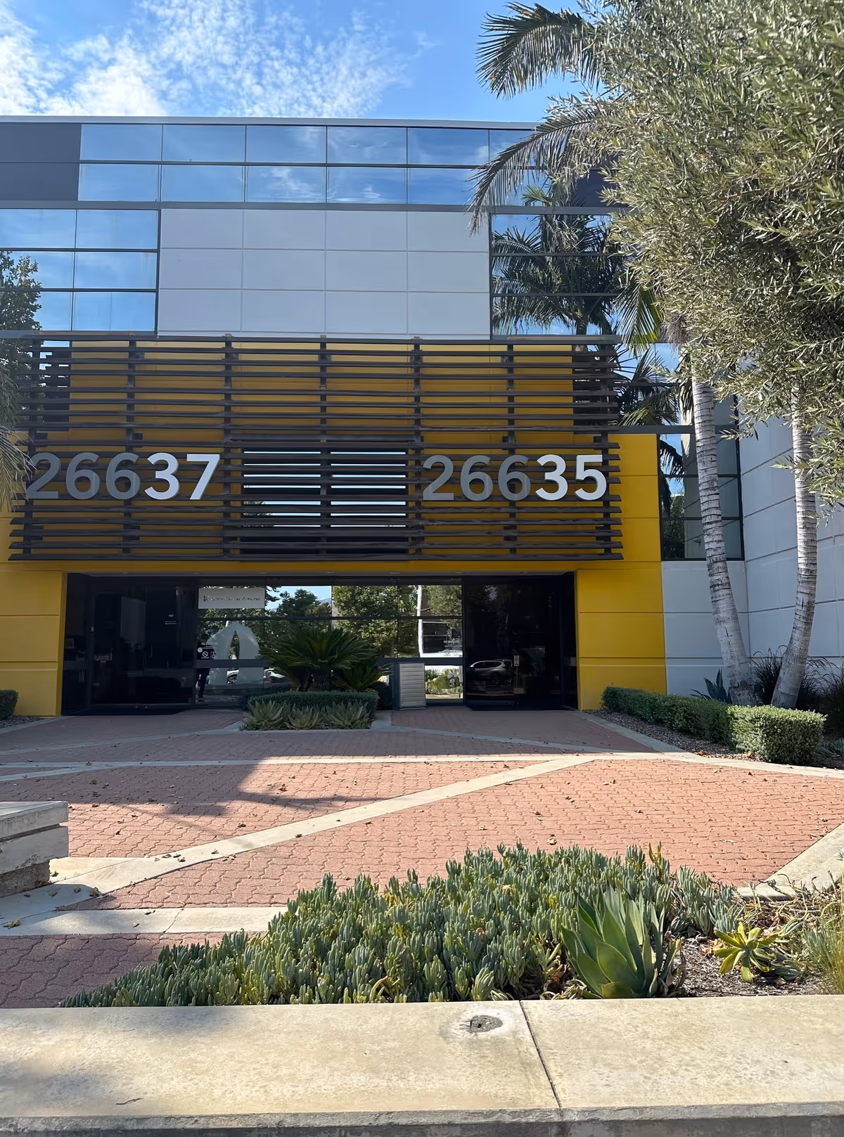 Exterior view of a modern building entrance with glass facade, yellow accents, and landscaping.