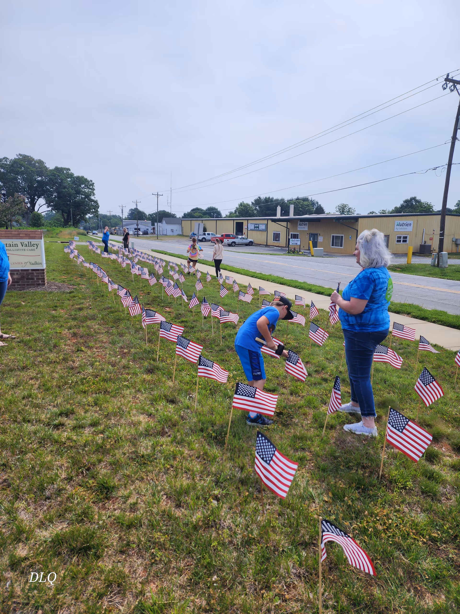 Grassy lawn with numerous American flags planted in rows, while people stand near a yellow building.