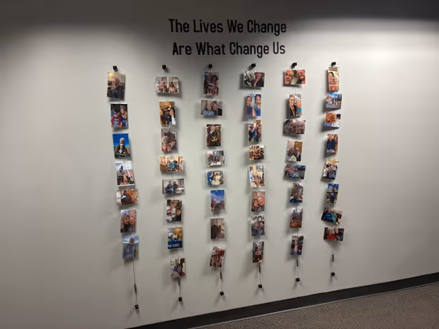 Interior wall with hanging photo strands under quote: 'The Lives We Change Are What Change Us'.