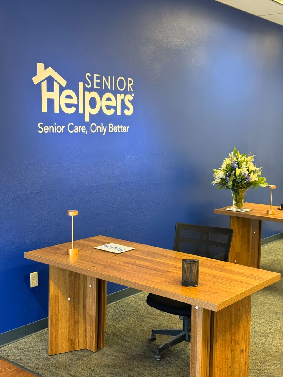Reception area with blue wall, Senior Helpers logo, wooden desk, chair, and flower bouquet.