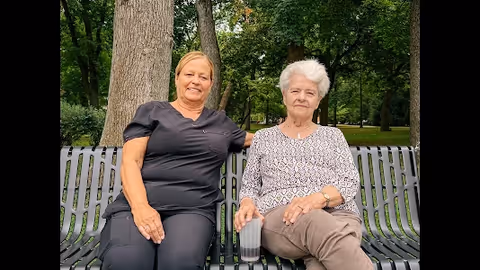 Two women sit on a metal bench in a park with trees.