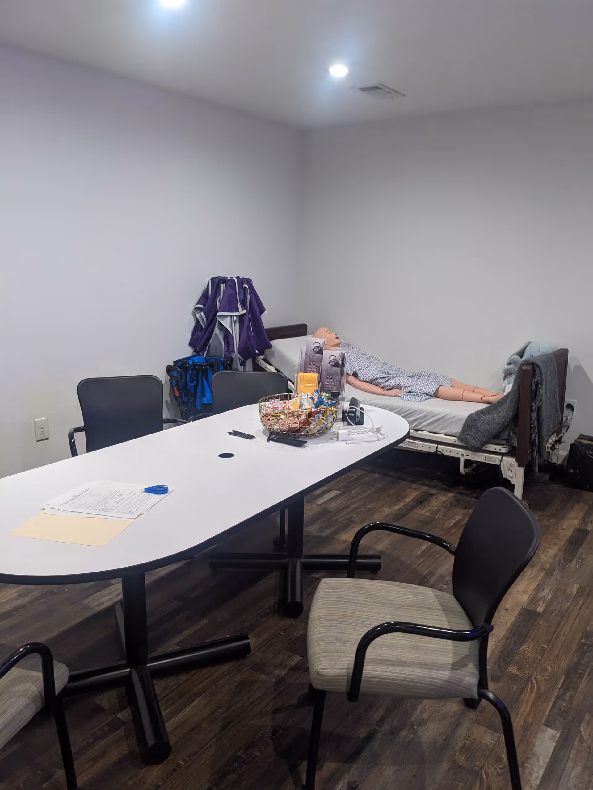 A small care room with a table, chairs, coats, and a bed at the back.