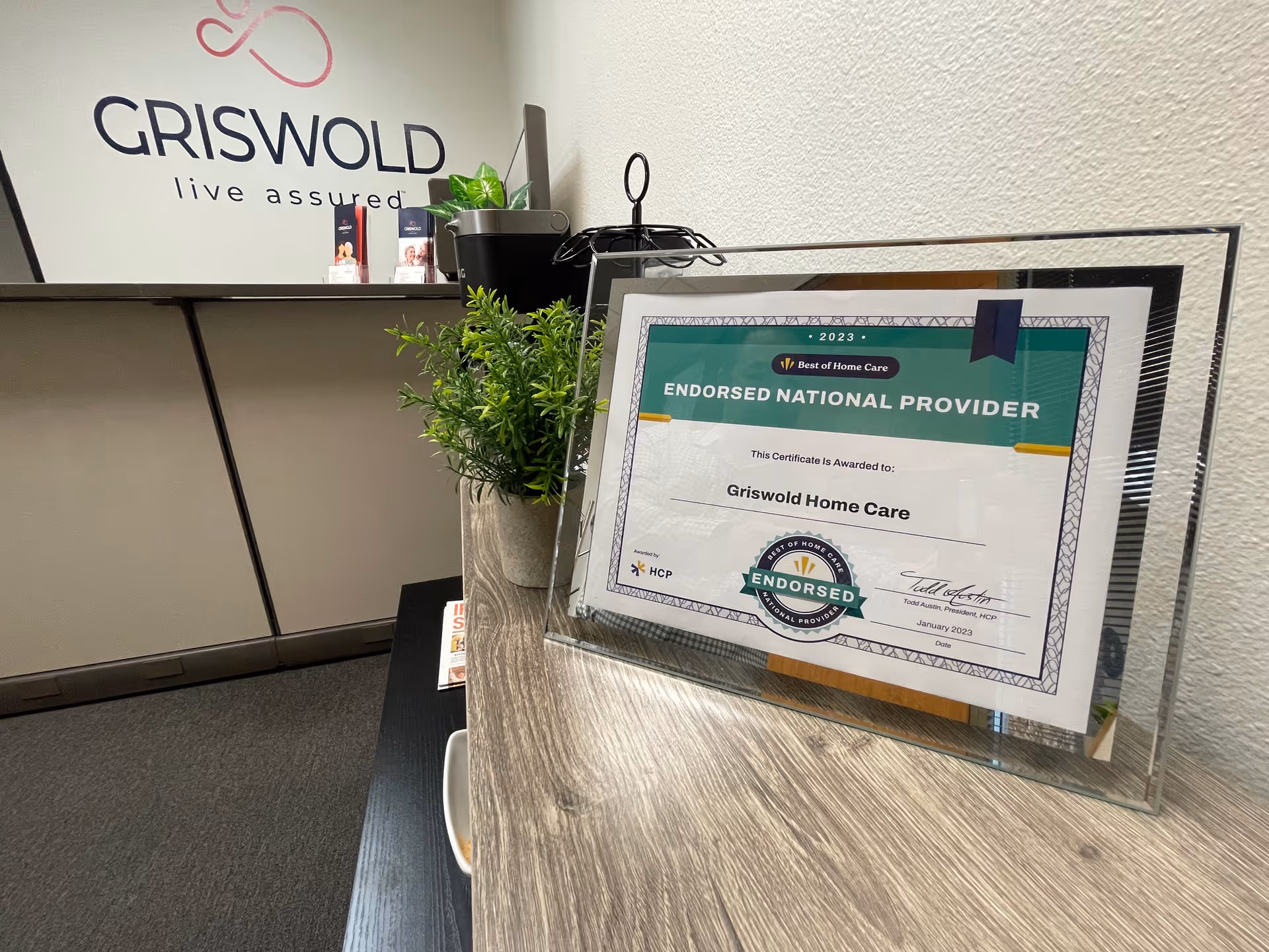 Framed certificate on a desk beside Griswold Home Care branding and a plant.