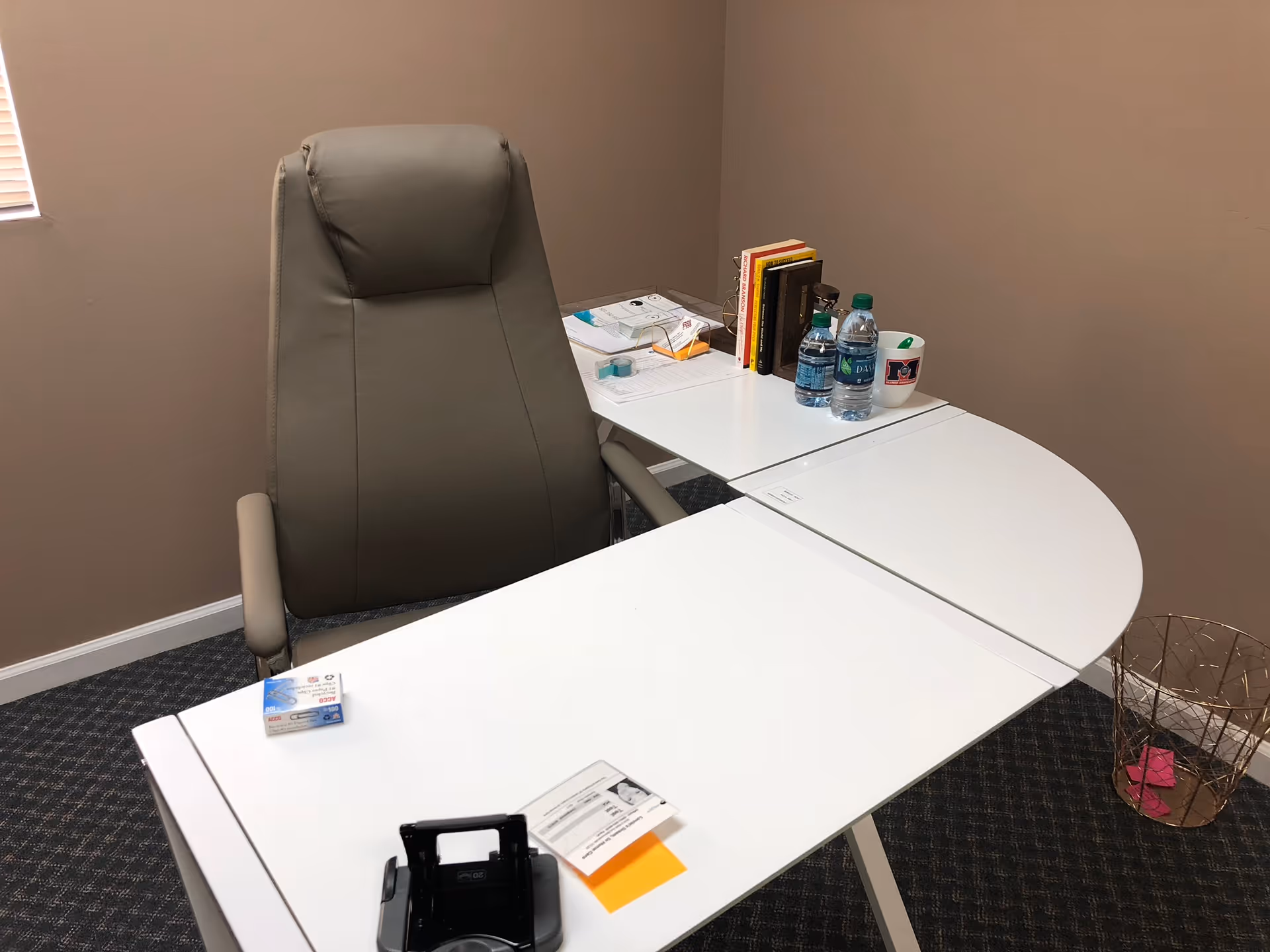 Curved white office desk with a gray chair, books, water bottles, and papers.