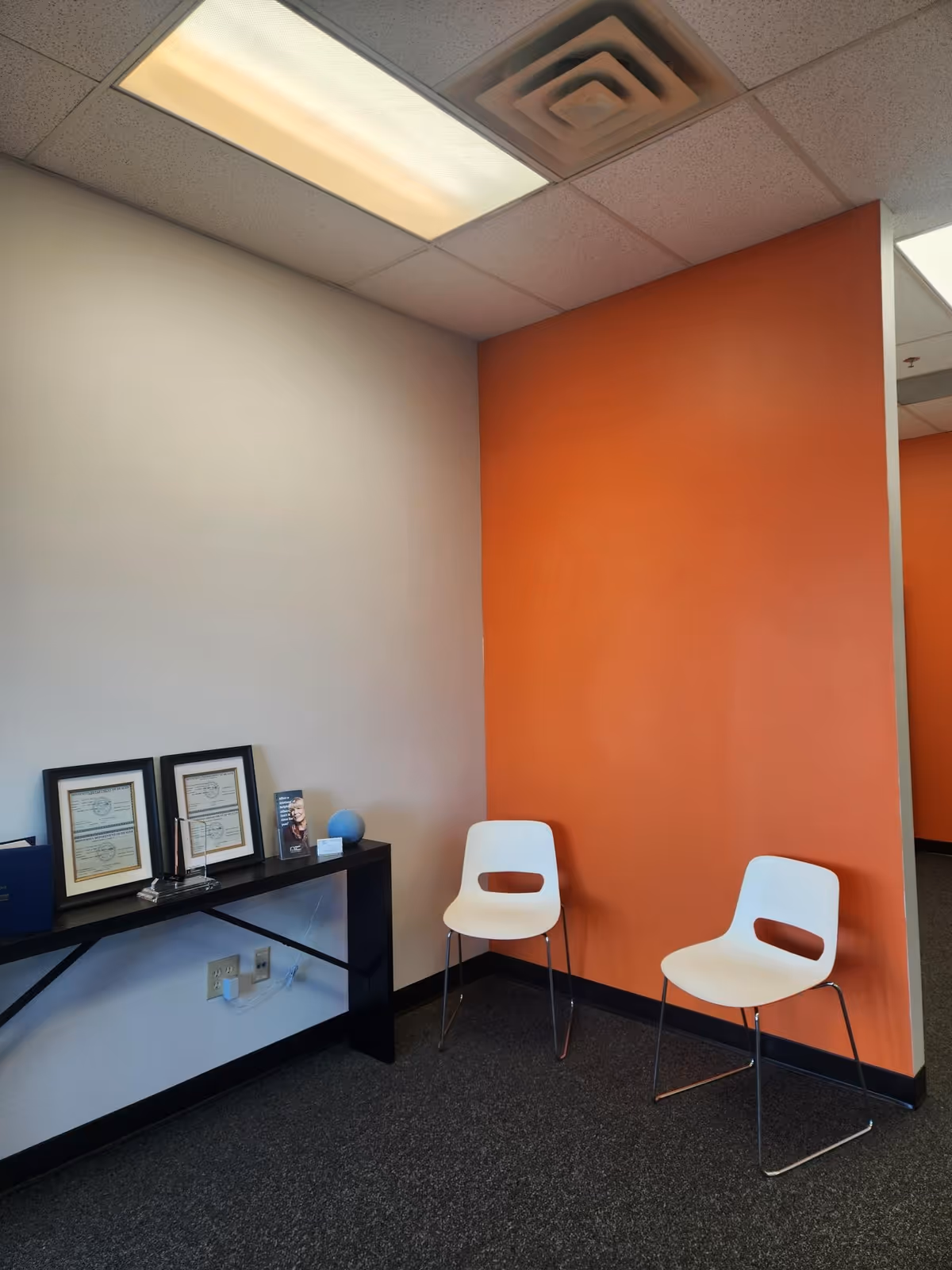 An interior waiting area with two white chairs and a black console against orange walls.