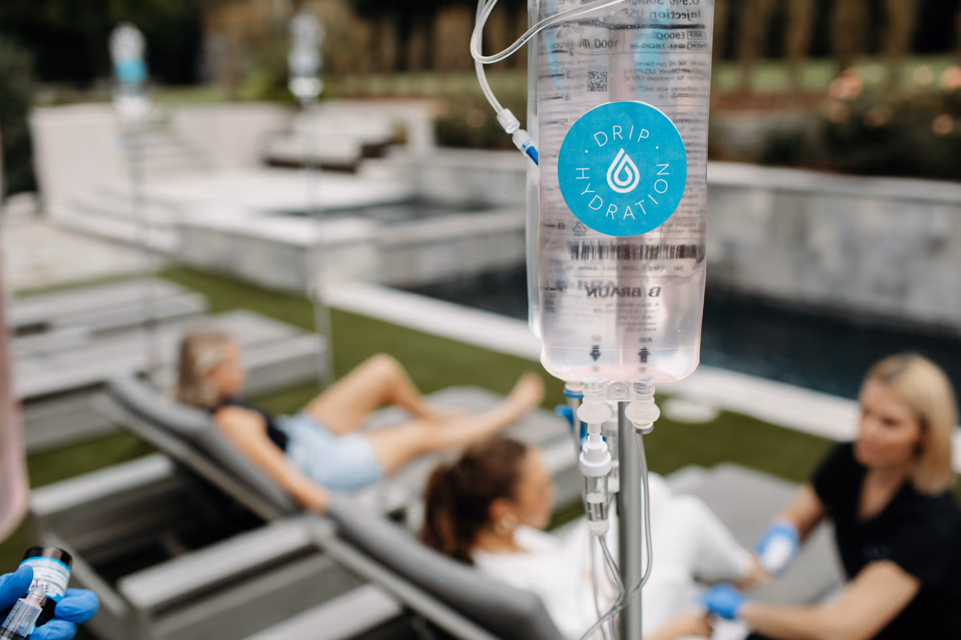 IV drip bag labeled Drip Hydration outdoors, with residents lounging by a poolside courtyard.