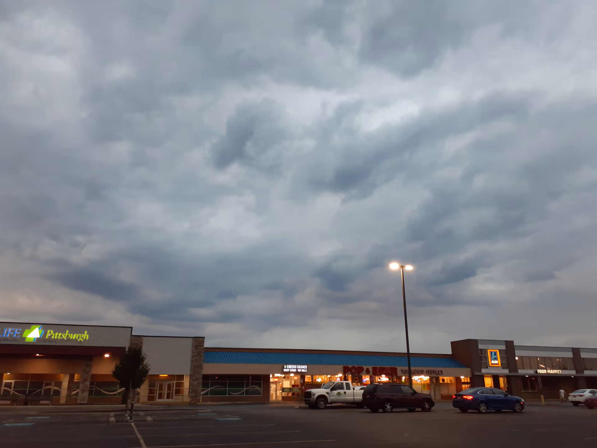 Overcast sky over a strip mall exterior with storefronts and parked cars.