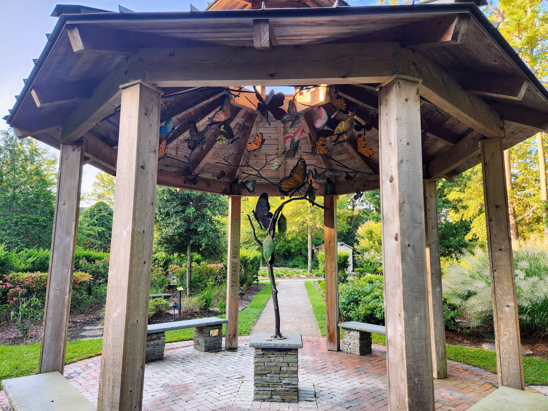 Wooden gazebo with butterfly sculptures in a brick courtyard amid lush garden.