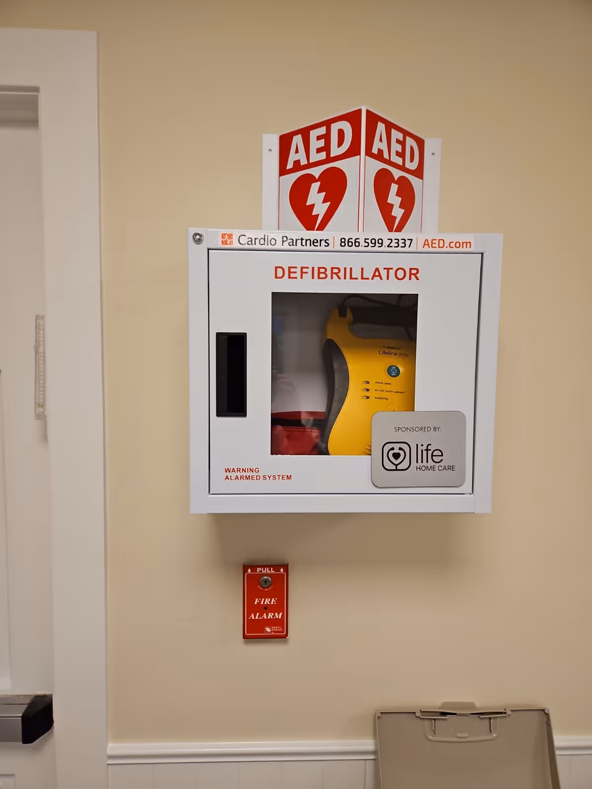 Wall-mounted AED defibrillator cabinet with sponsor label in a hallway