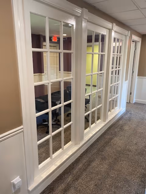 Interior hallway with white-framed glass partition windows and seating beyond, carpeted corridor.