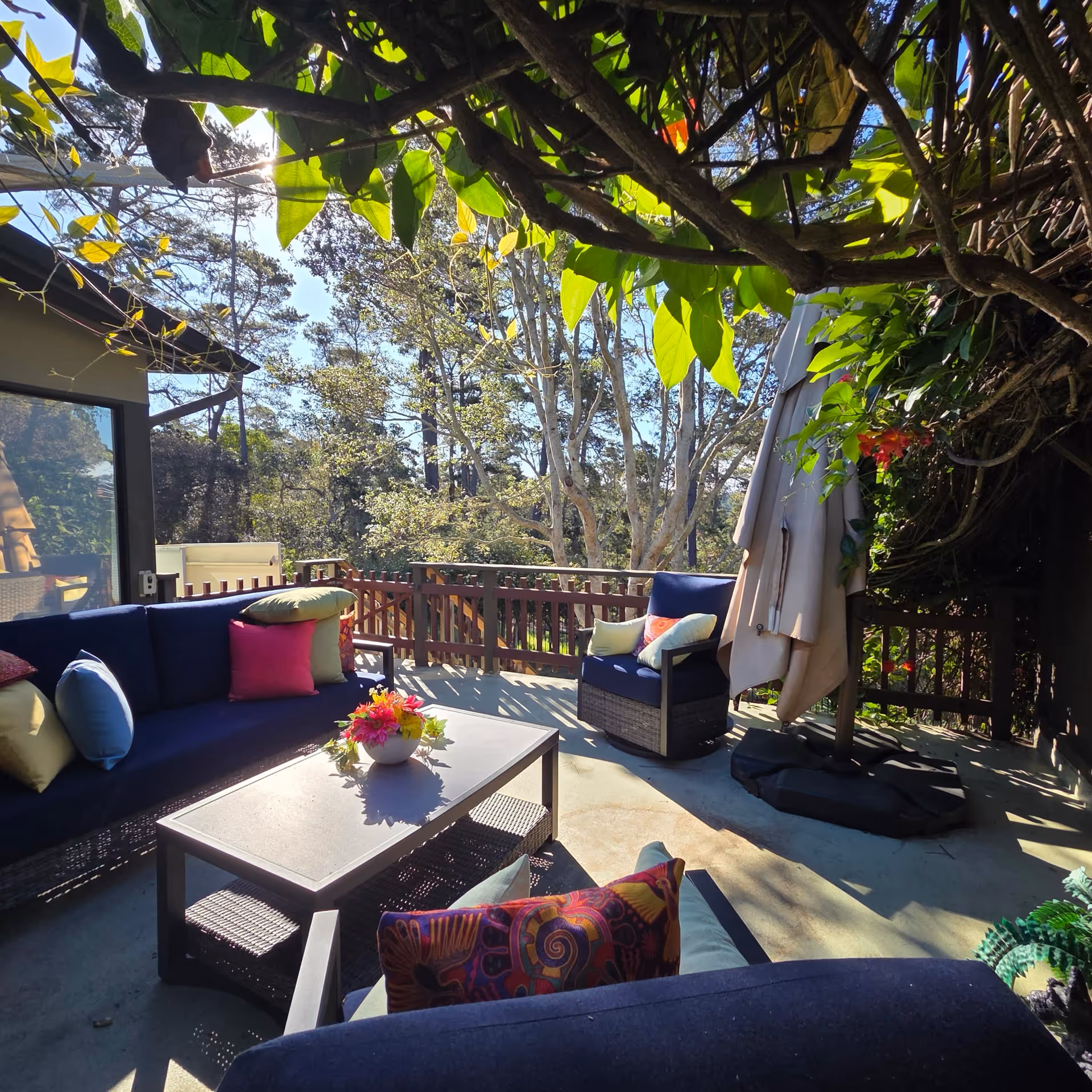 Sunny outdoor balcony with blue cushioned seating, colorful pillows, and a coffee table.