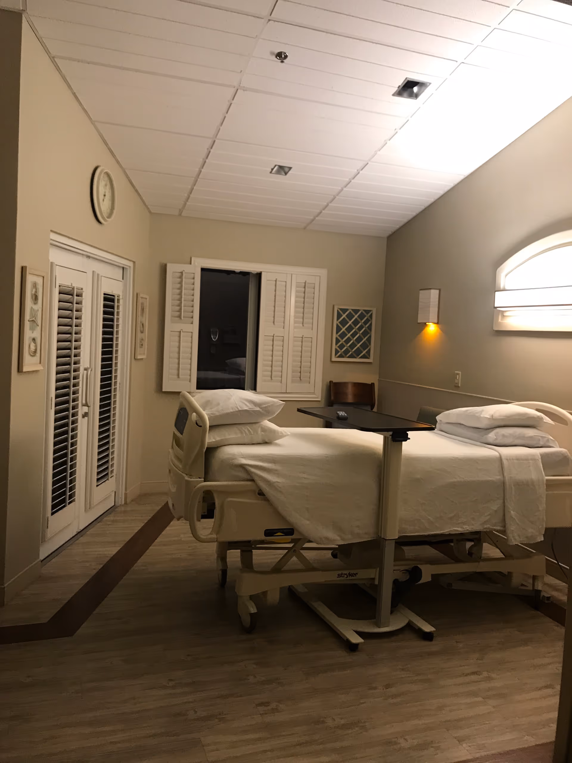 Two hospital beds with white linens in a softly lit patient room with window shutters.