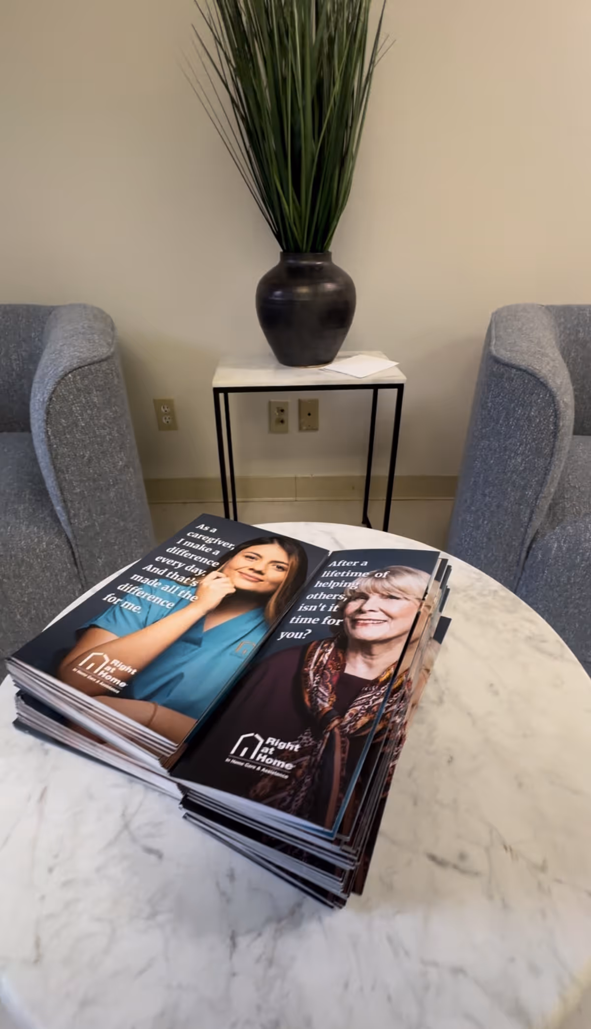 Two gray lounge chairs face a marble table with brochures and a tall plant.