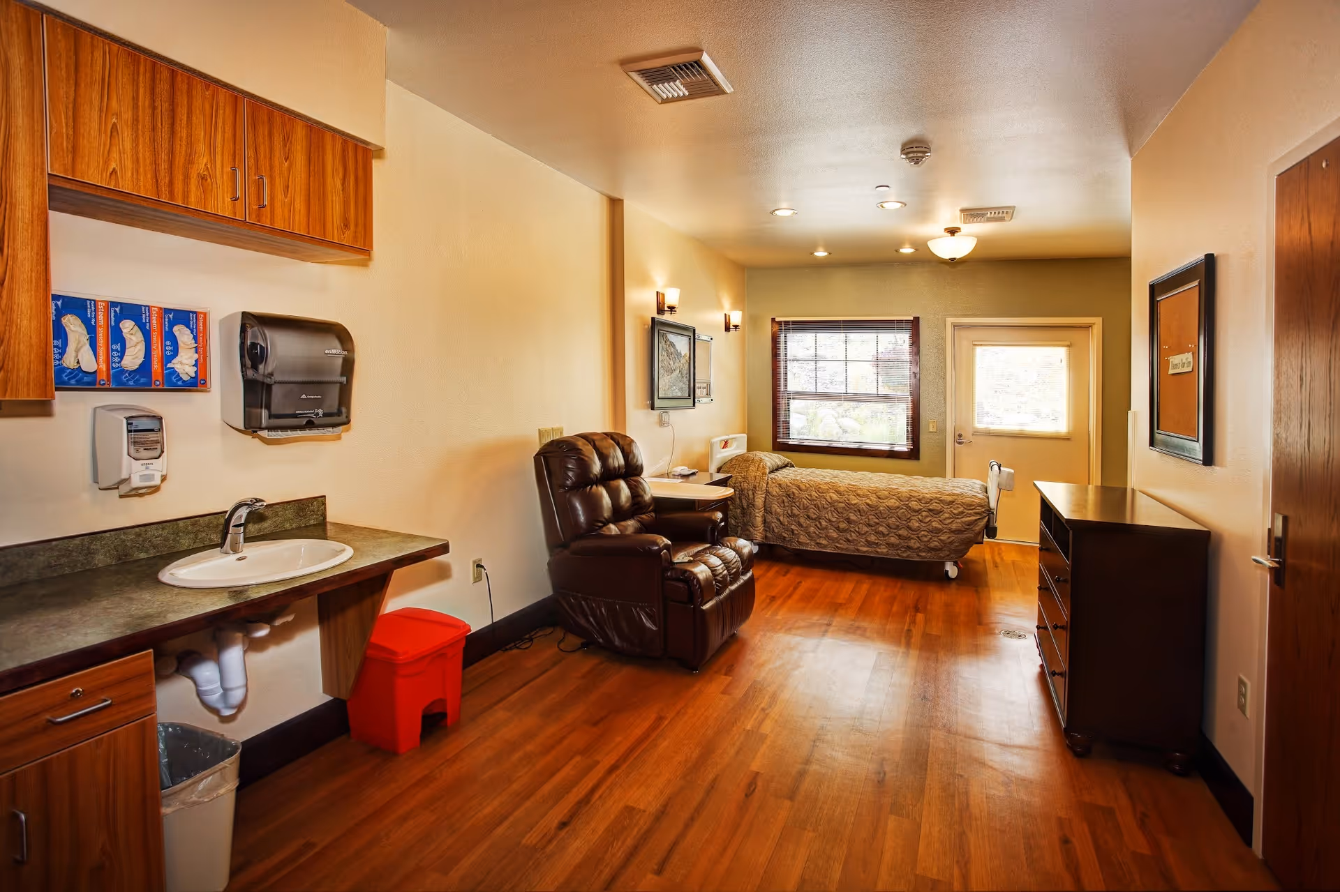 Hospice living area with a bed, recliner, kitchenette, and dresser in a sunny room.
