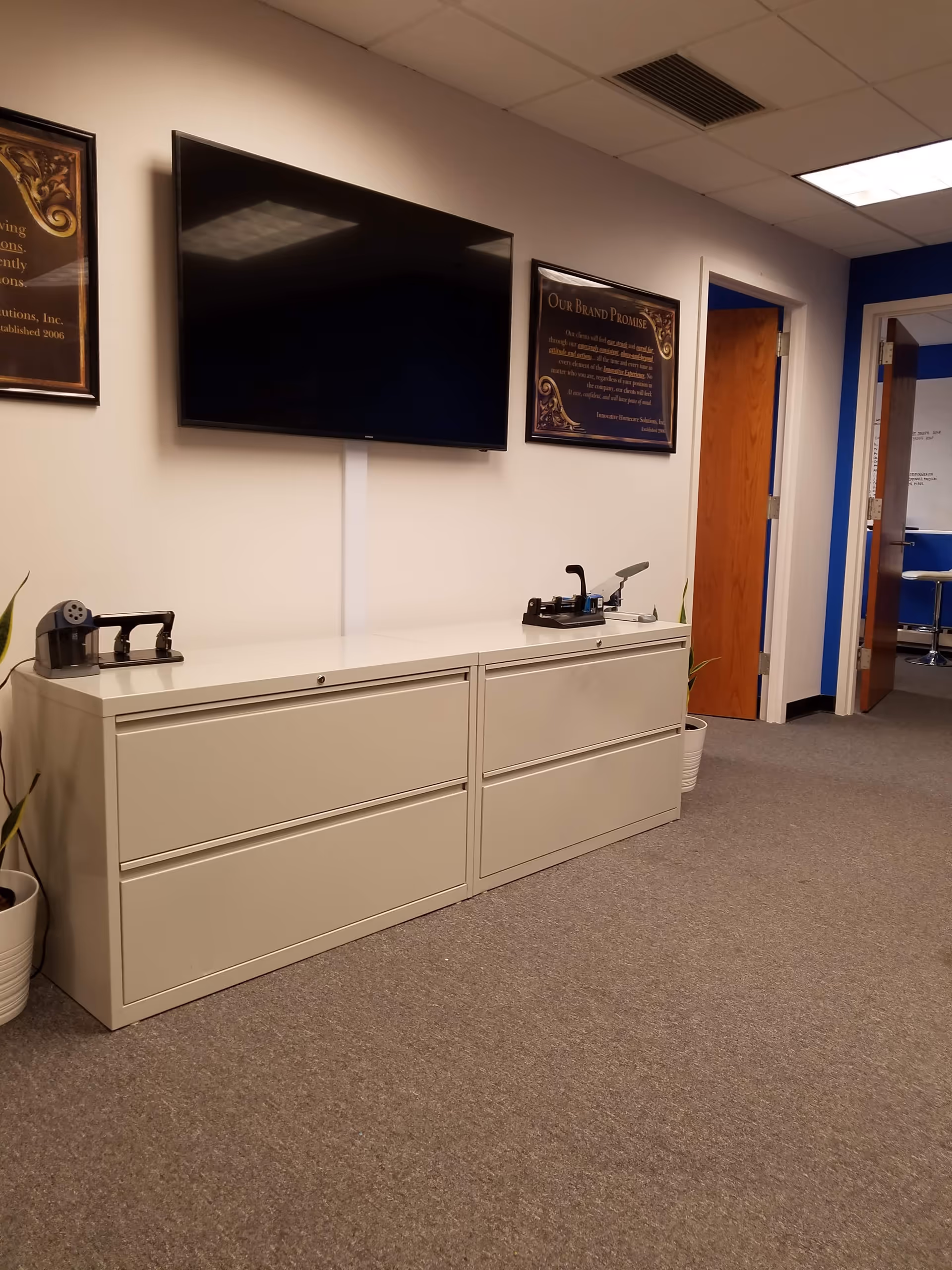 Interior office area with a wall-mounted TV, beige filing cabinets, and framed plaques.