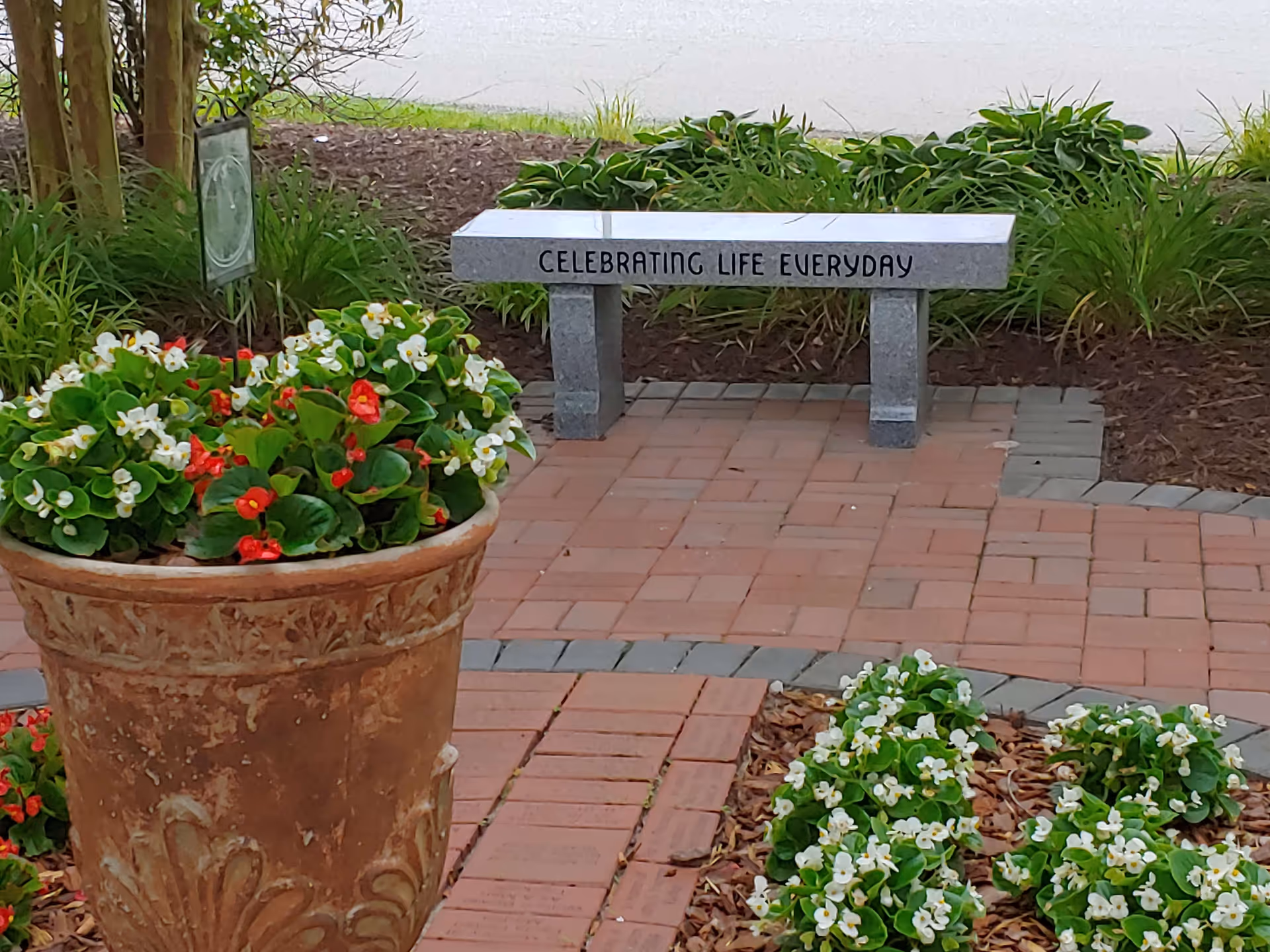 Outdoor brick walkway with flower planters and a bench inscribed Celebrating Life Everyday.