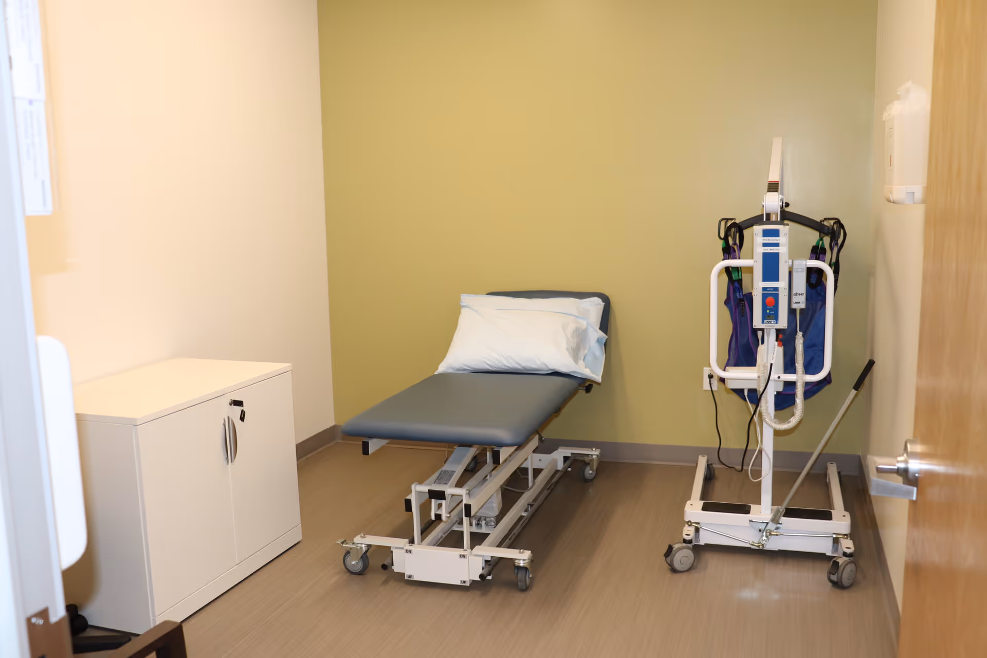 Medical examination room with a padded exam table, cabinet, and tall medical equipment on wheels.