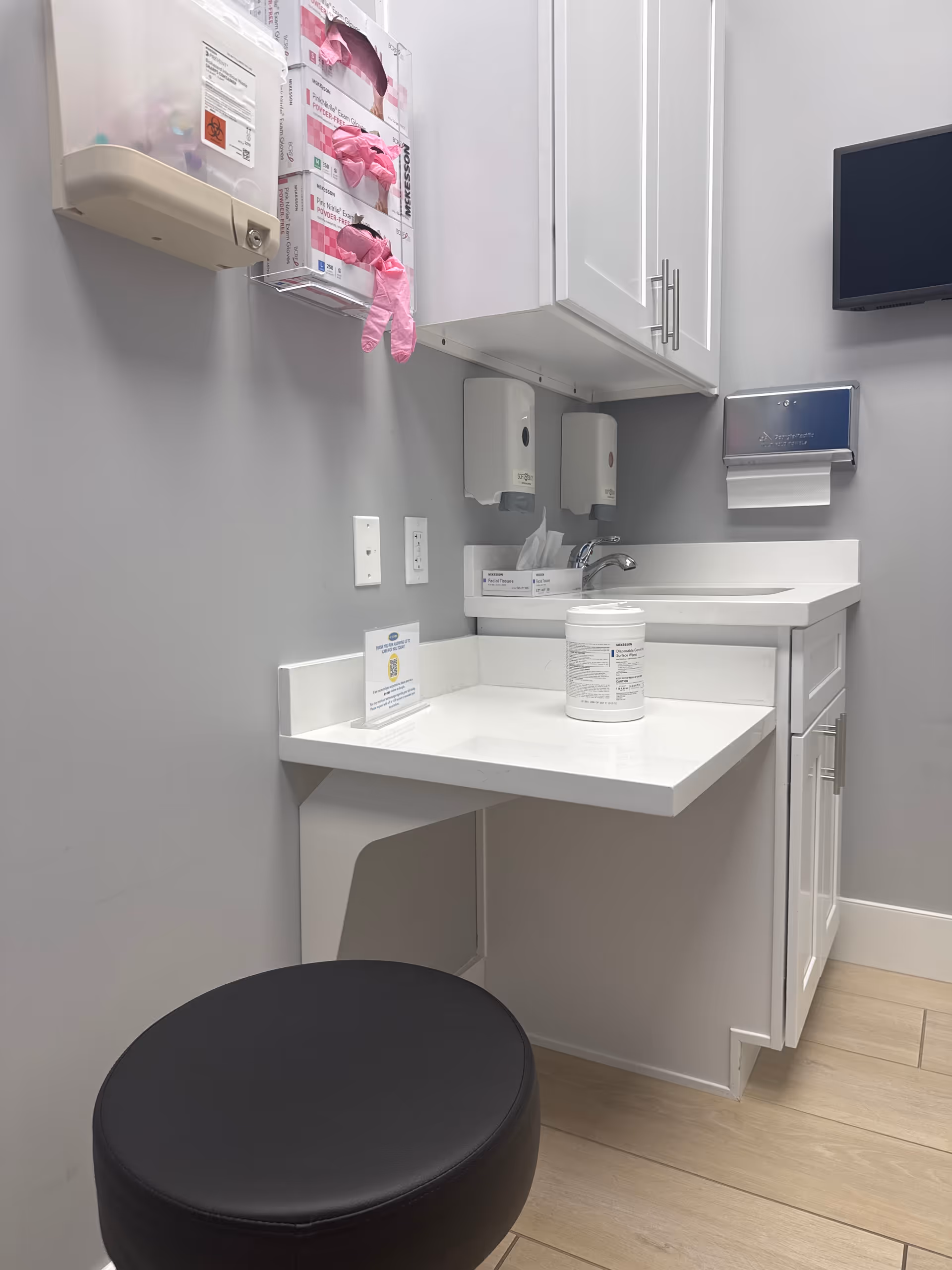 Clinical sink area with white cabinets, hand sanitizers, and a black round stool.
