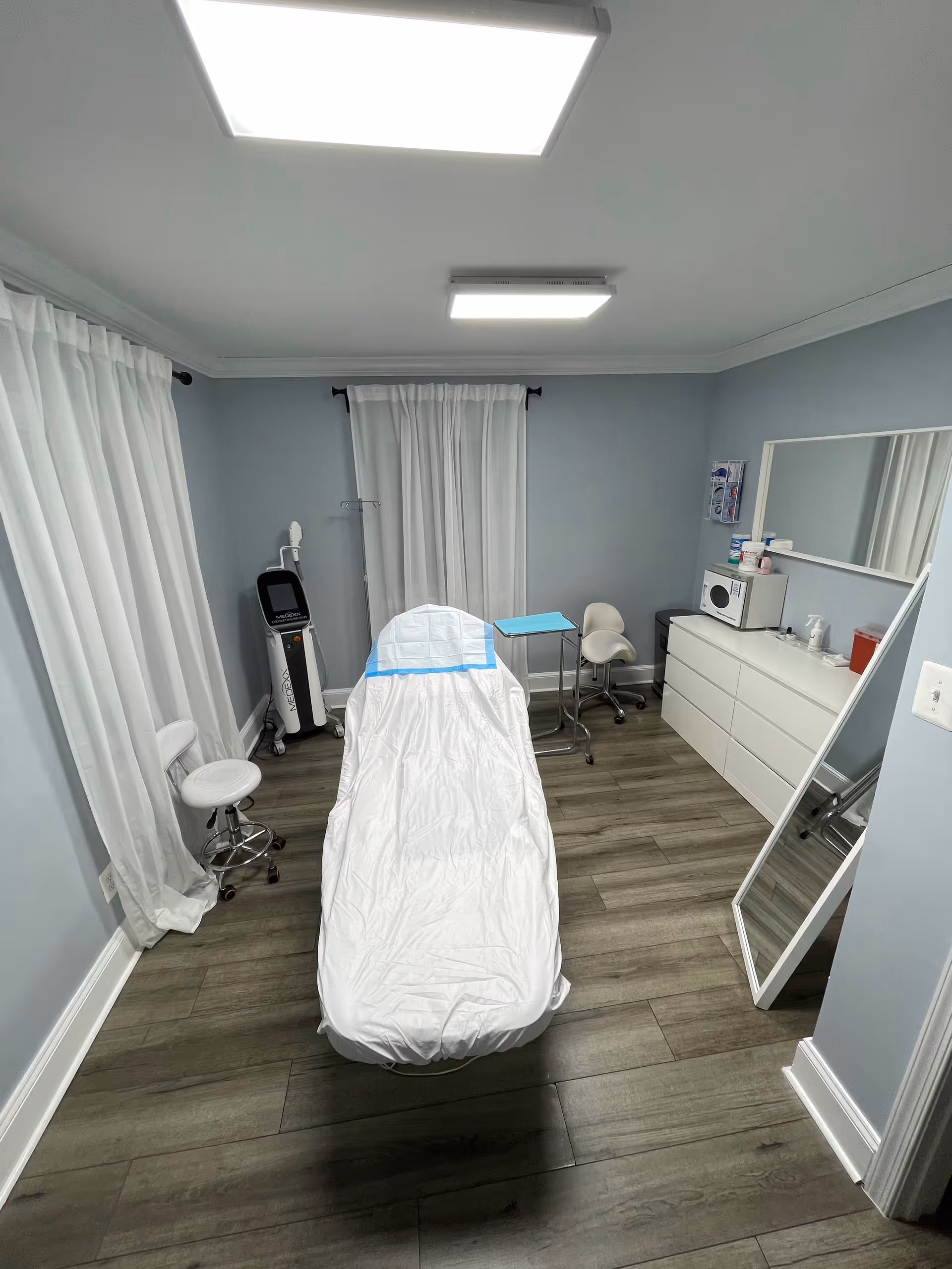 Treatment room with a covered bed, privacy curtains, and medical equipment against soft blue walls.