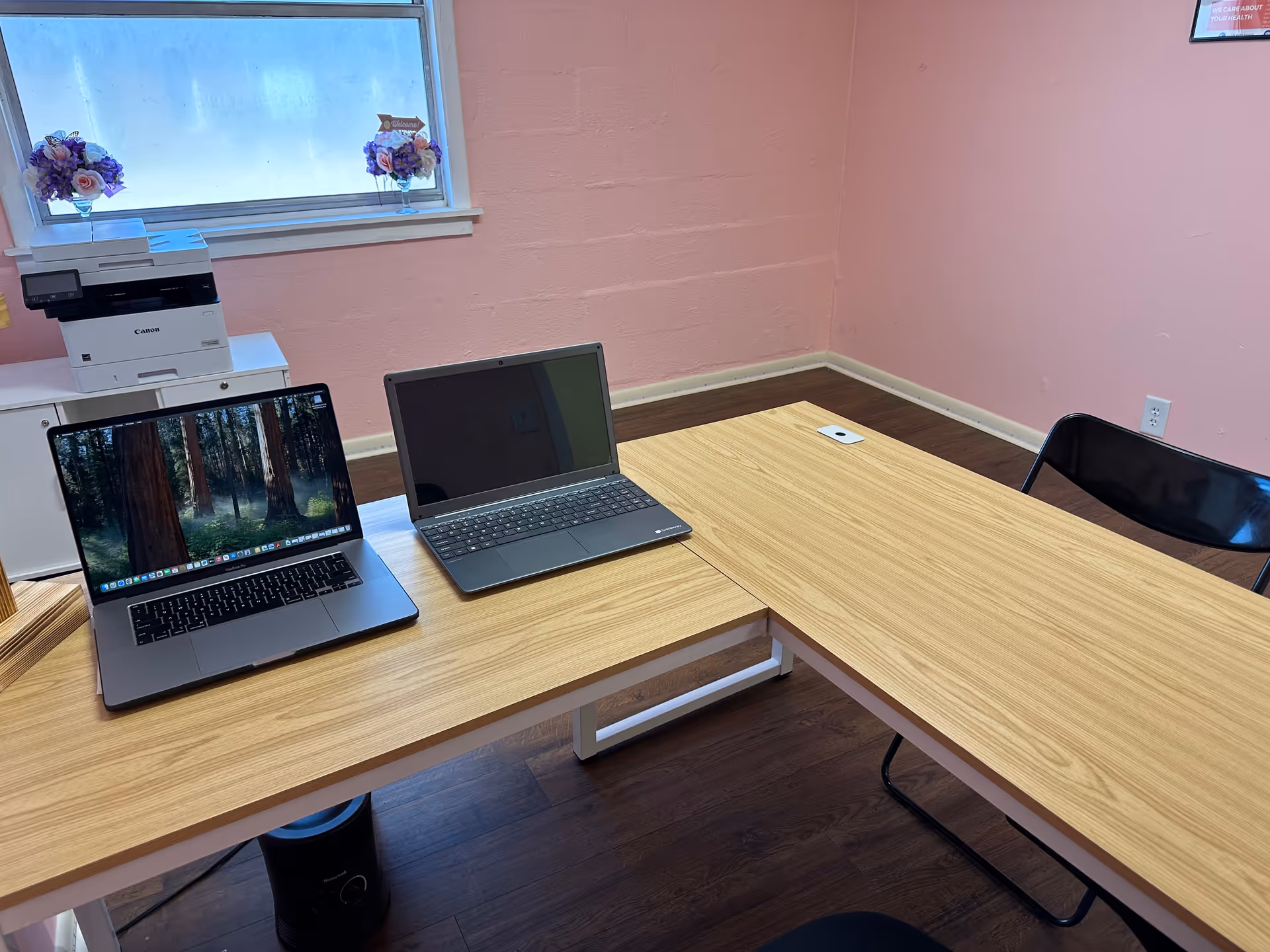 Pink-walled office area with two laptops, a printer, and a window with flowers.