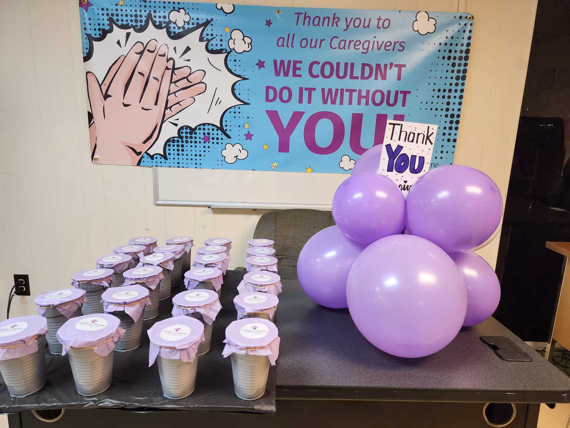 Indoor event setup with balloons and cups on a table, banner thanking caregivers.