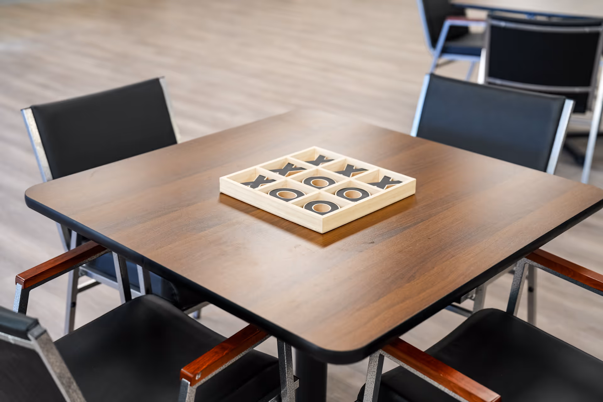 Wooden dining table with a tic tac toe board and black chairs in a common area.