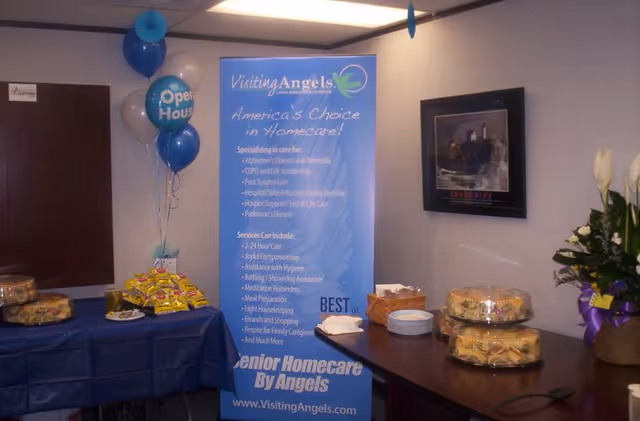 Indoor promotional setup with a blue Visiting Angels banner, balloons, and snack table.