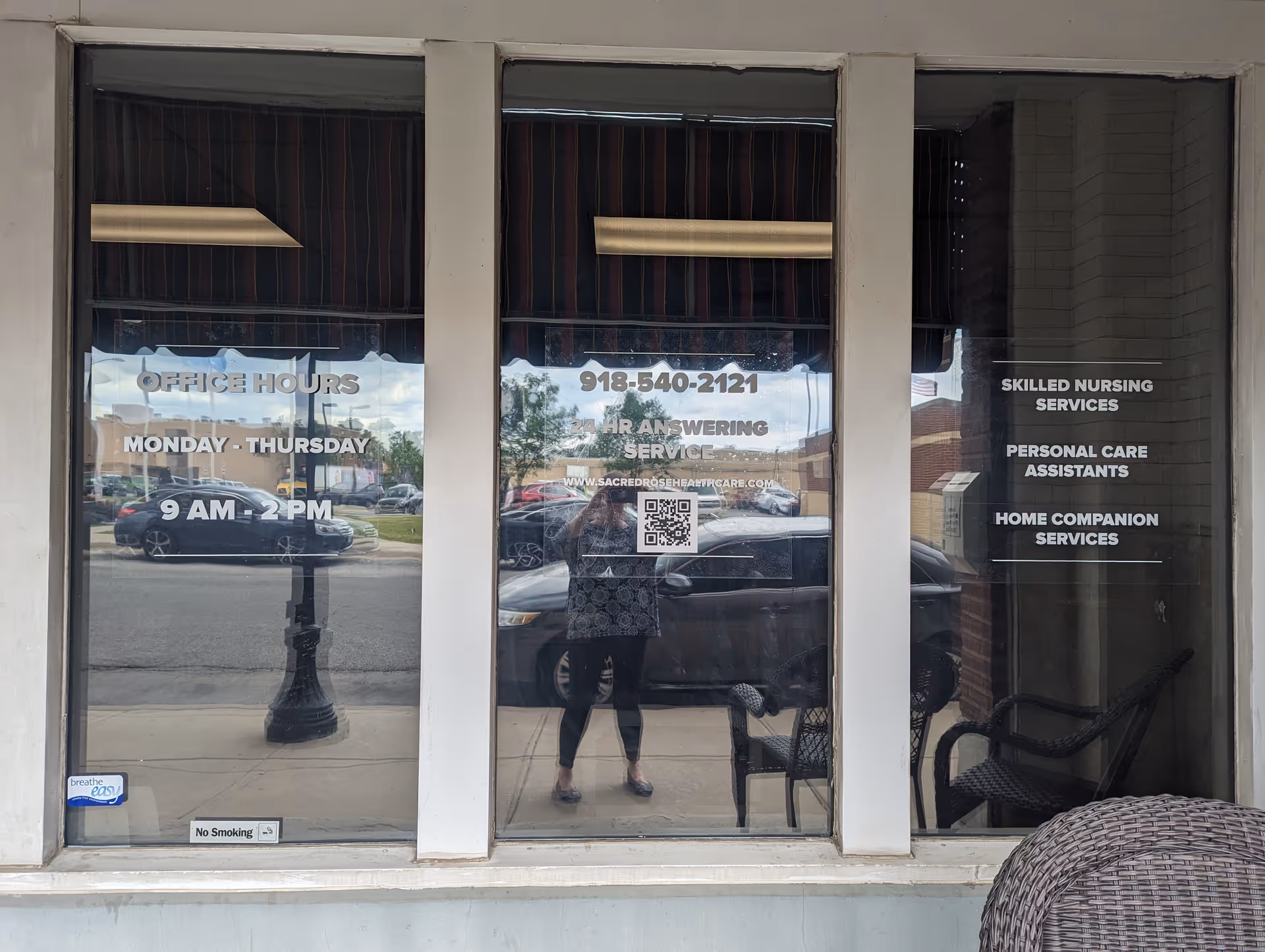 Storefront window for Sacred Rose Healthcare displaying hours and services; a person is reflected.