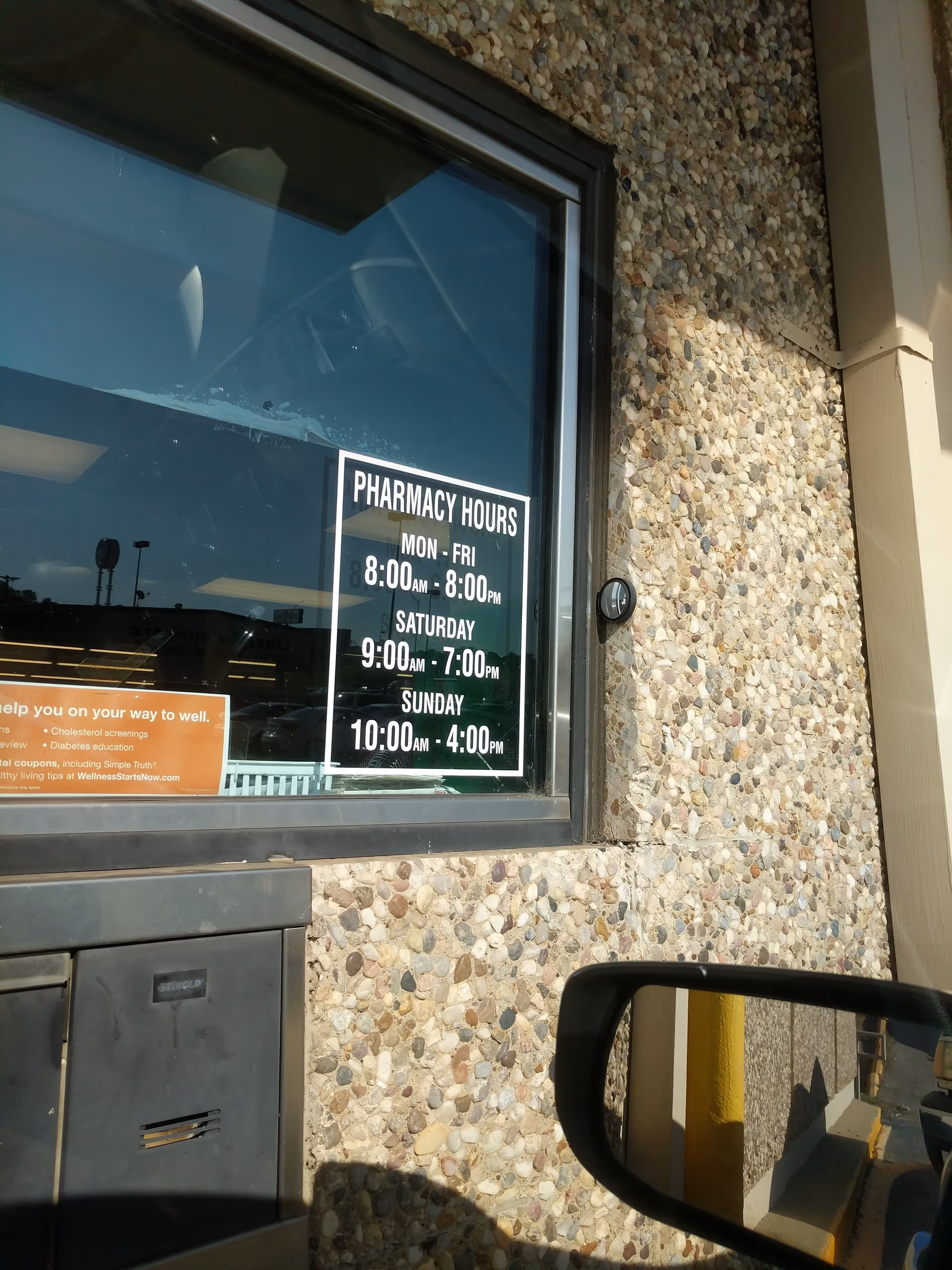 Exterior wall with a window displaying pharmacy hours and a car side mirror.