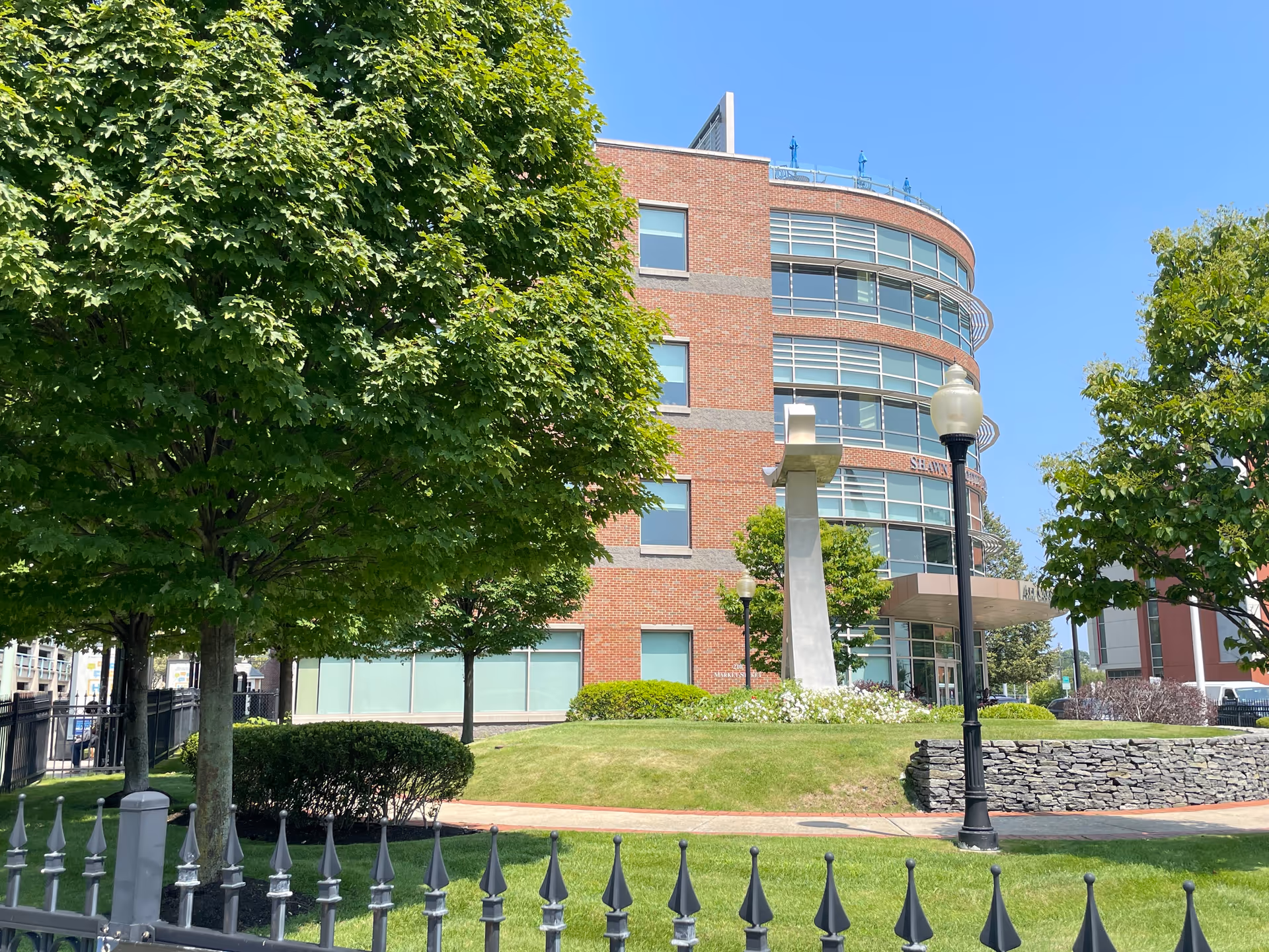 Brick senior living building with curved glass windows, green lawn, trees, and street lamps.