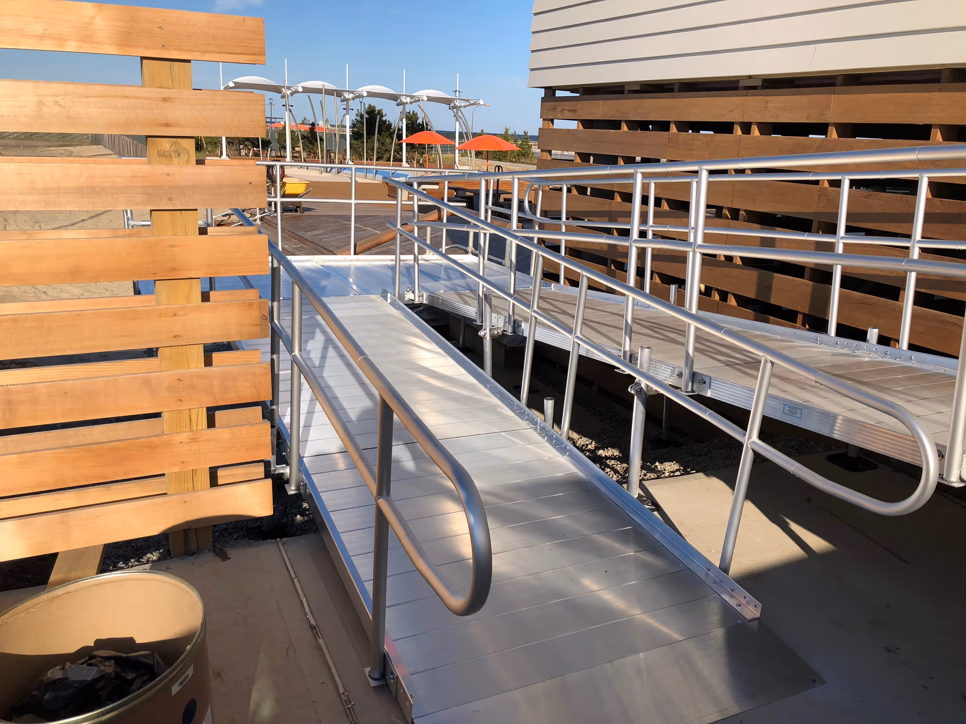 Outdoor metal ramp with handrails leads to a wooden-sided facility, with lounge umbrellas in distance.