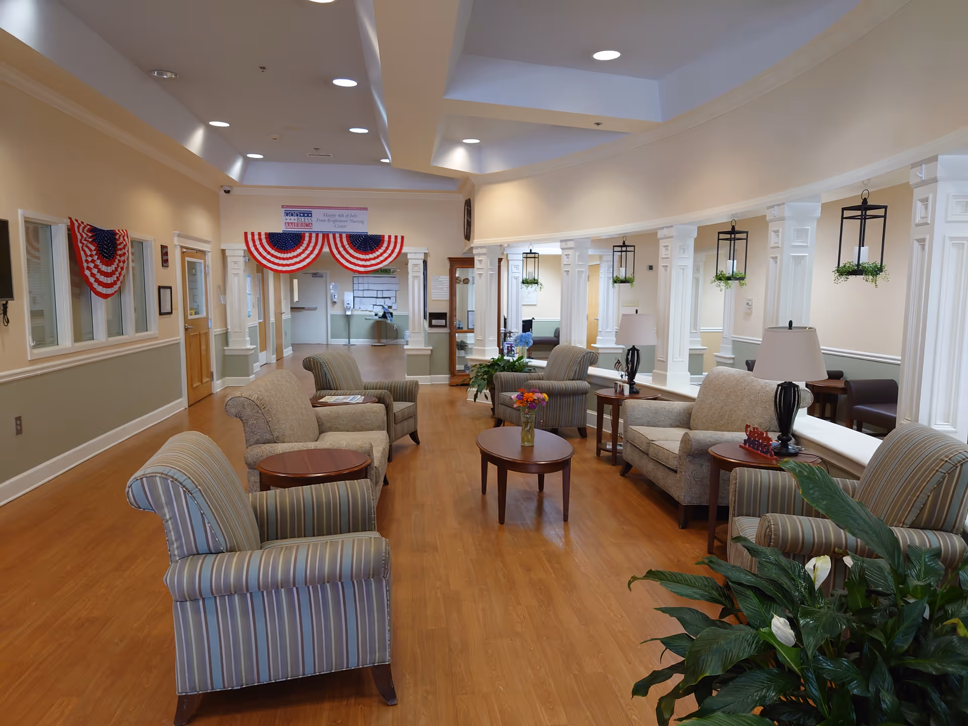 Bright, spacious senior living lounge with arranged seating, tables, plants, and patriotic banners.