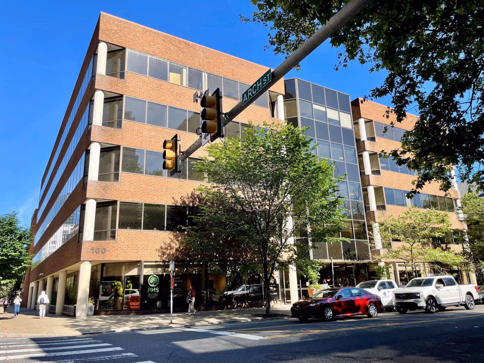 Brick and glass office building on a sunny city street with trees and cars.
