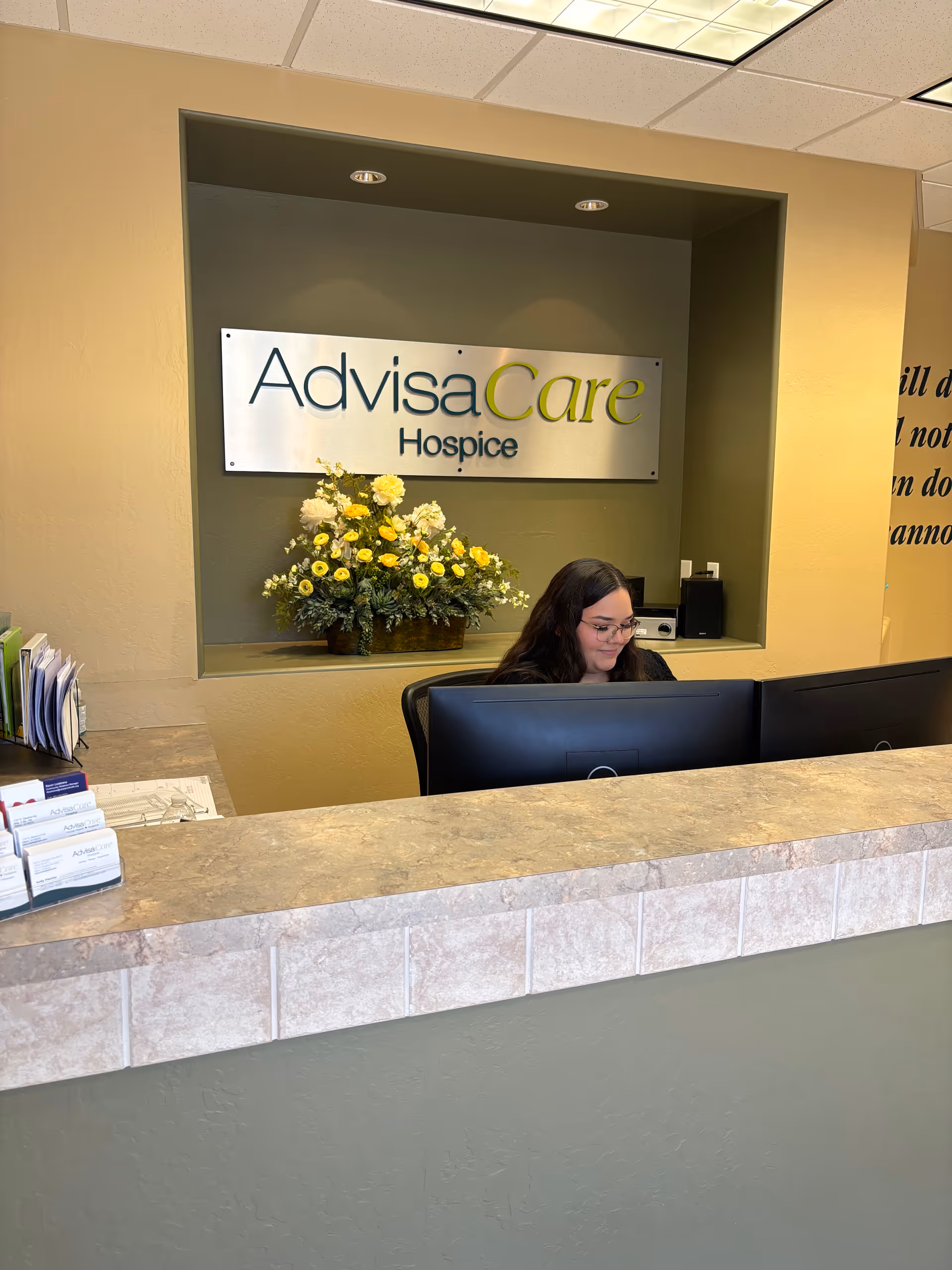 Reception desk with staff member, AdvisaCare Hospice sign, and flowers in a lobby.