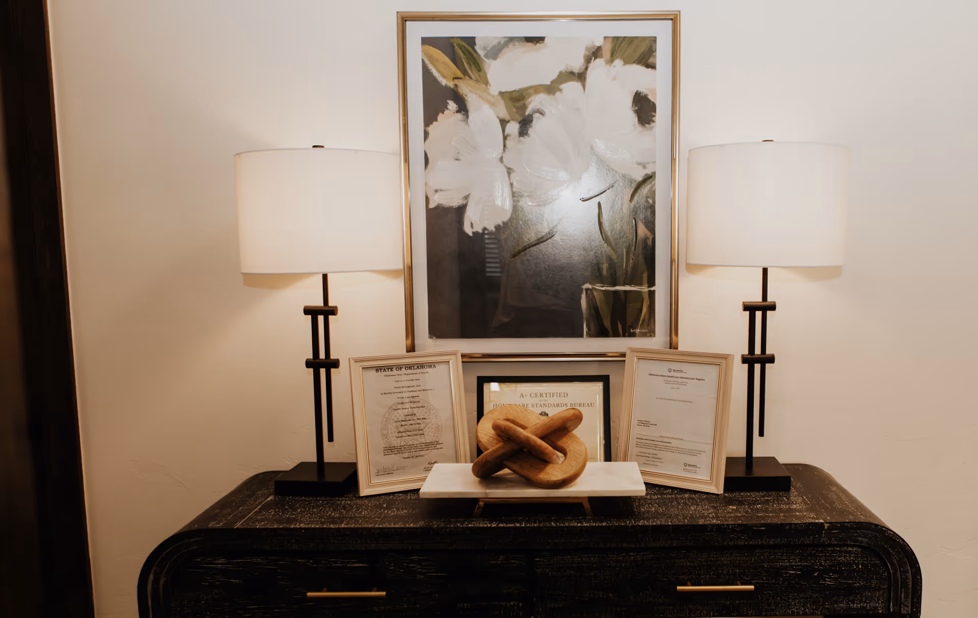 A dark wooden console with two lamps, framed certificates, a painting, and a decorative knot sculpture.