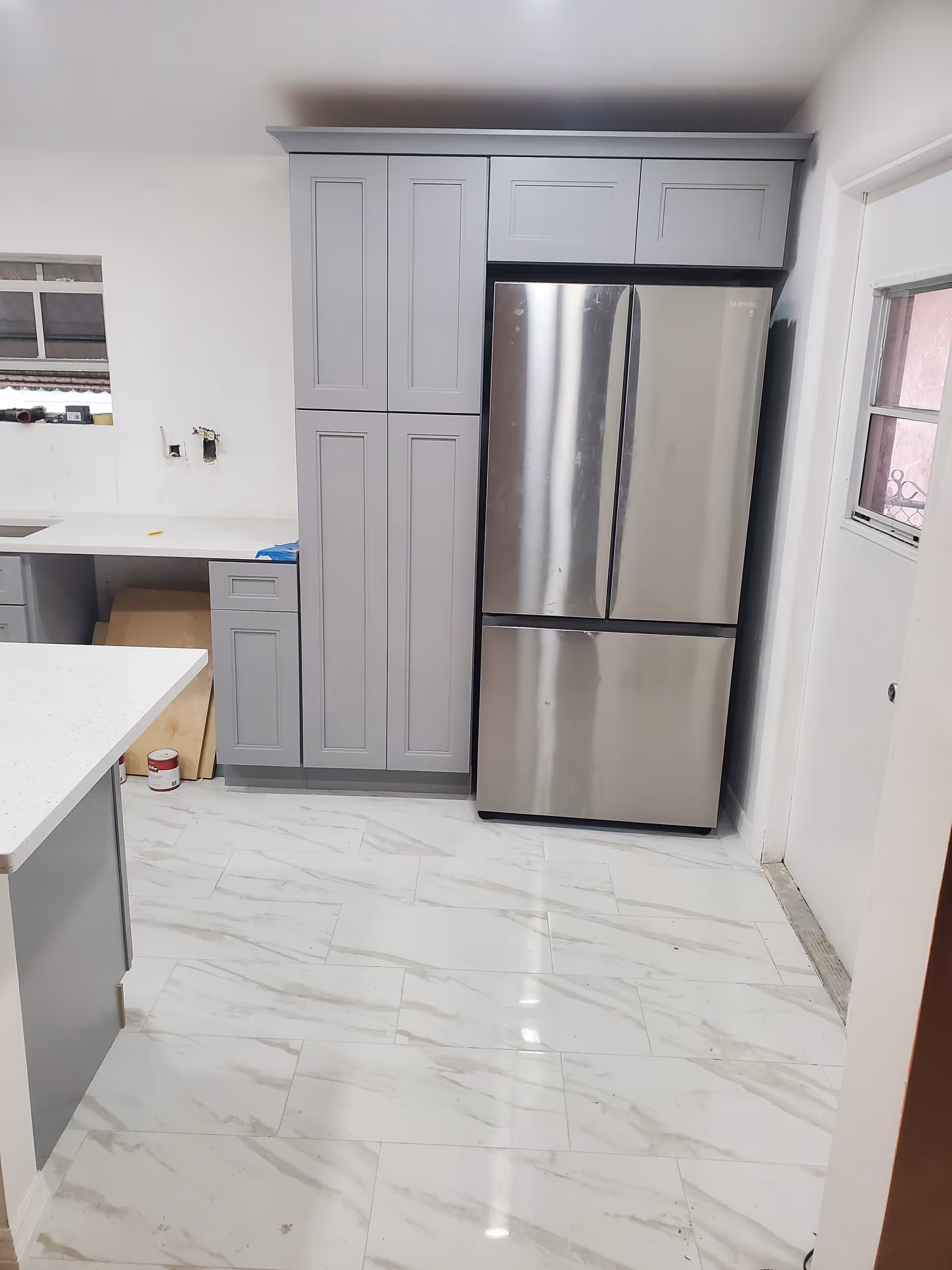 Modern kitchen with gray cabinets, stainless steel refrigerator, white countertops, and marble-look floor.